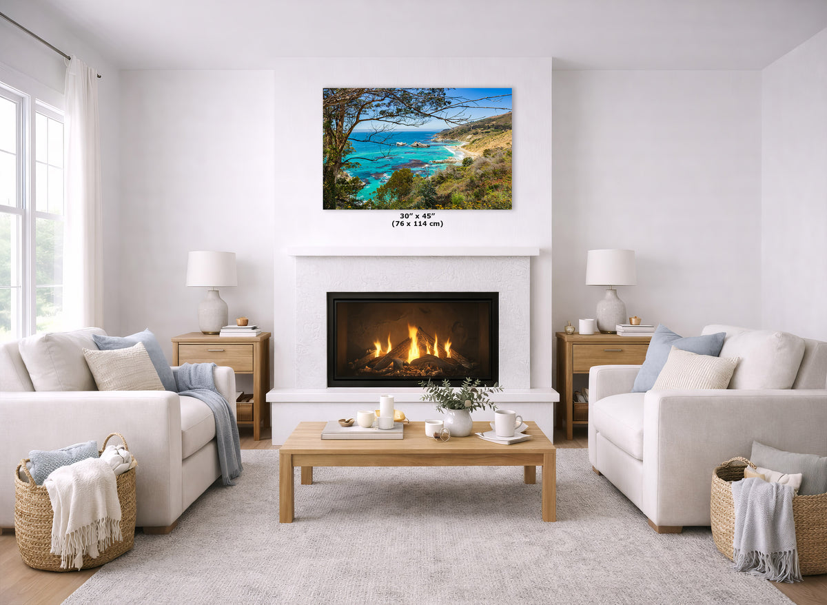 A coastal landscape wall art print hangs above a fireplace in a bright, modern living room. The artwork shows turquoise ocean water, a sandy beach, and green hills under a blue sky.