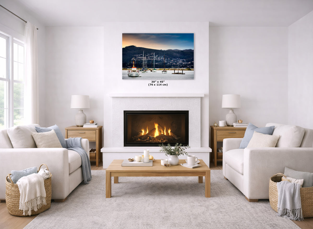 A landscape art print depicting a harbor town at sunset is displayed on a white wall above a fireplace in a bright, modern living room with two white sofas and a wooden coffee table.