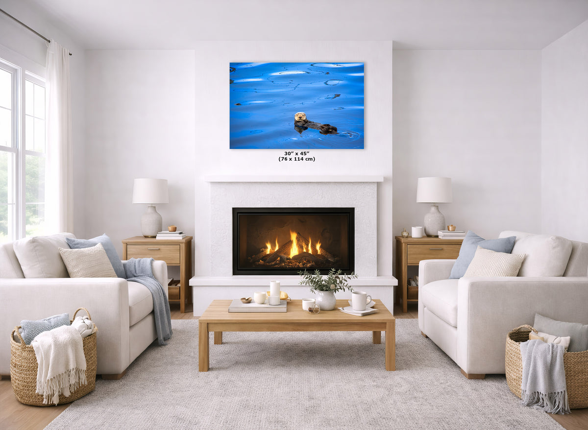 A canvas print of a blue water scene with a small otter floating in the water, displayed above a white fireplace in a bright, modern living room with two white sofas and a wooden coffee table.