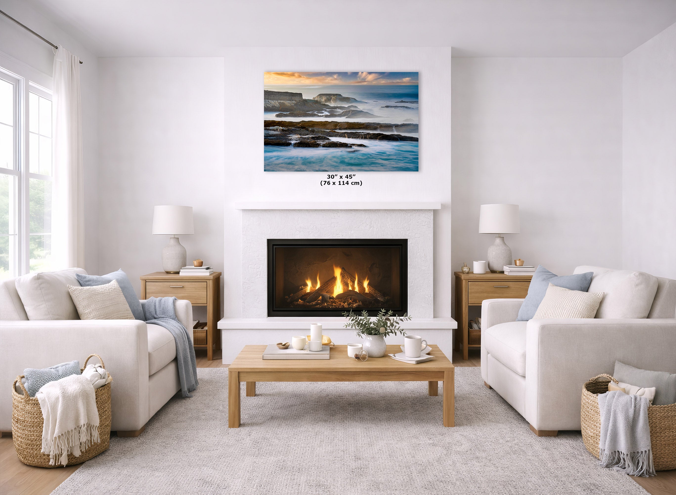 A landscape art print of a rocky coastline at sunset hangs above a white fireplace in a bright, modern living room with two cream sofas, a wooden coffee table, and a light rug.