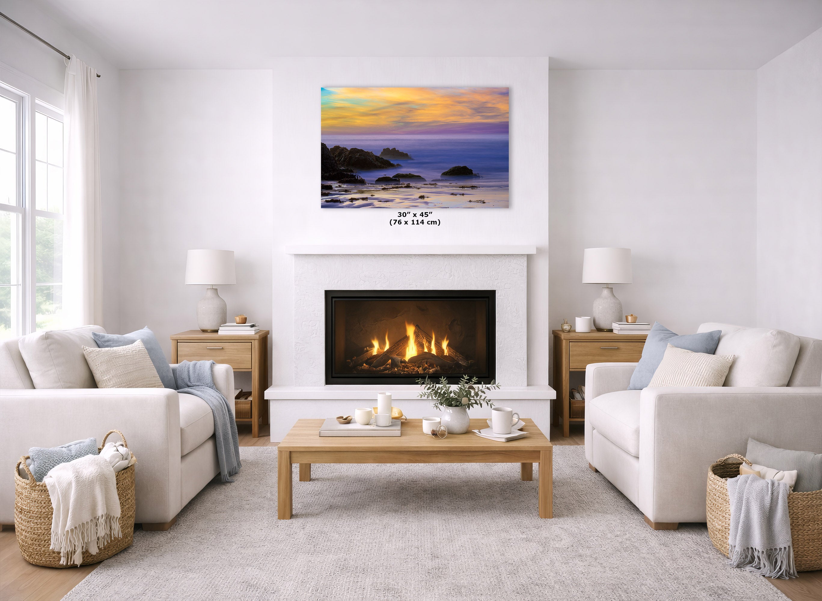 A framed coastal sunset wall art print is displayed above a white fireplace in a bright, modern living room with two cream sofas, a wooden coffee table, and a light rug. The print shows a beach and ocean at sunset with blue, purple, orange, and yellow tones.
