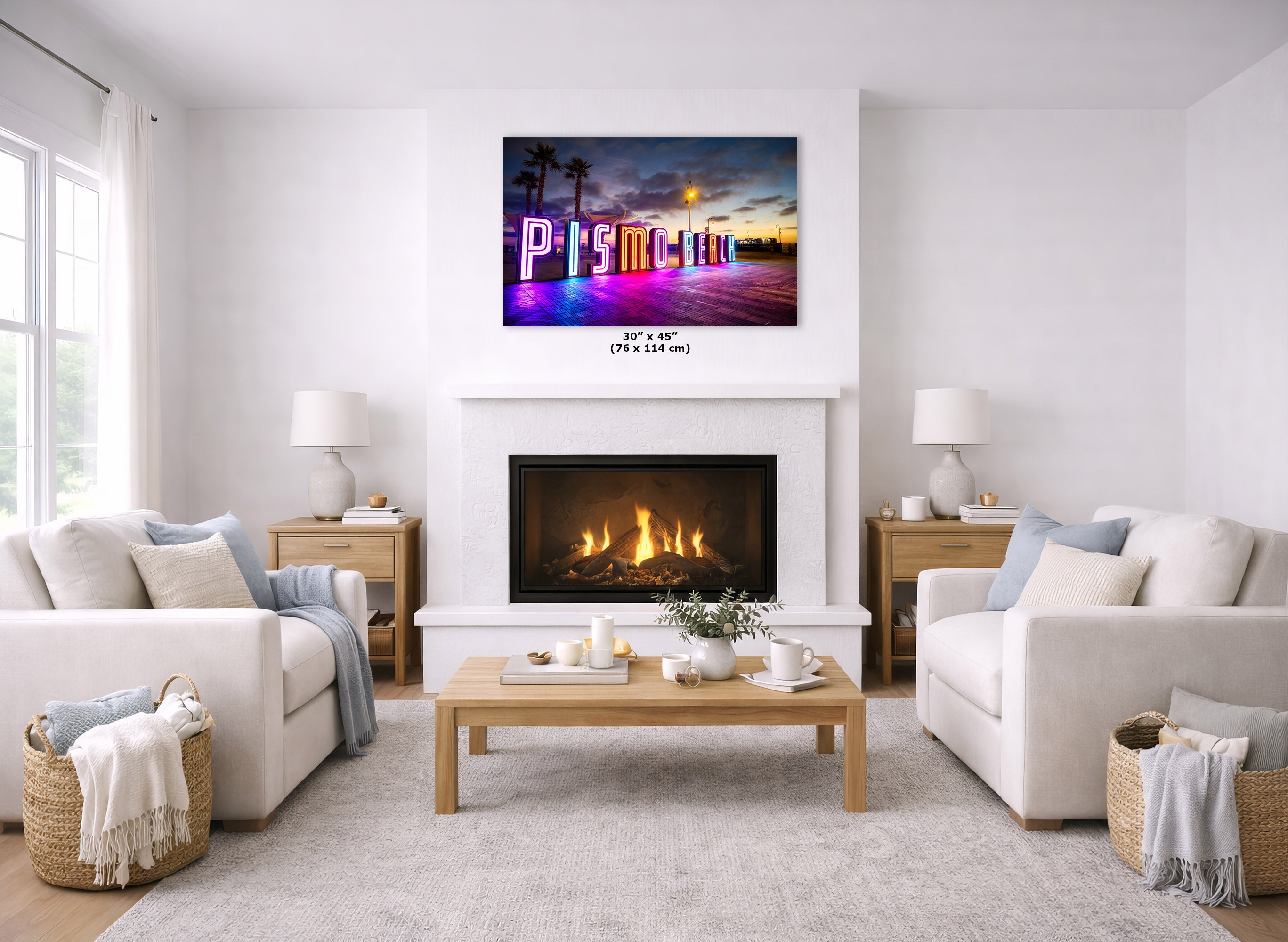A wall art print showing a colorful beach scene with large letters spelling 'Pismo' is displayed above a fireplace in a bright, modern living room with two white sofas and a wooden coffee table.
