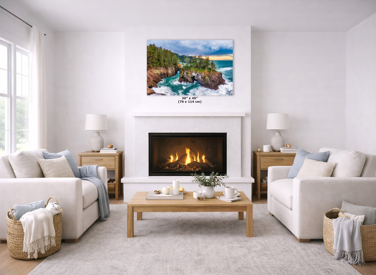 A coastal landscape art print hangs above a white fireplace in a bright, modern living room with two cream sofas, a wooden coffee table, and a light rug. The print depicts turquoise water, rocky cliffs, and green trees under a blue sky.