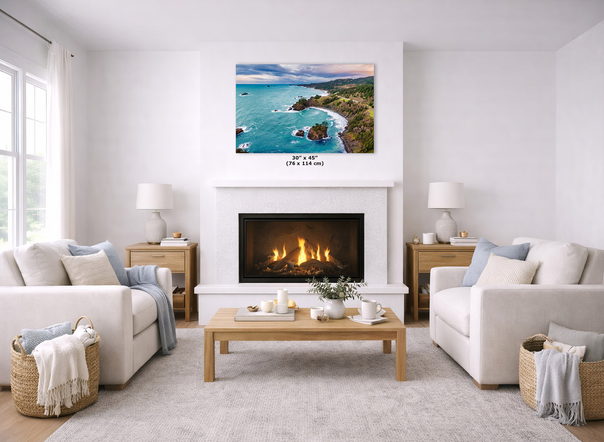 A coastal landscape wall art print hangs above a white fireplace in a bright, modern living room with two cream sofas, a wooden coffee table, and a light rug. The print shows turquoise ocean water, rocky coastline, and a sunset sky.