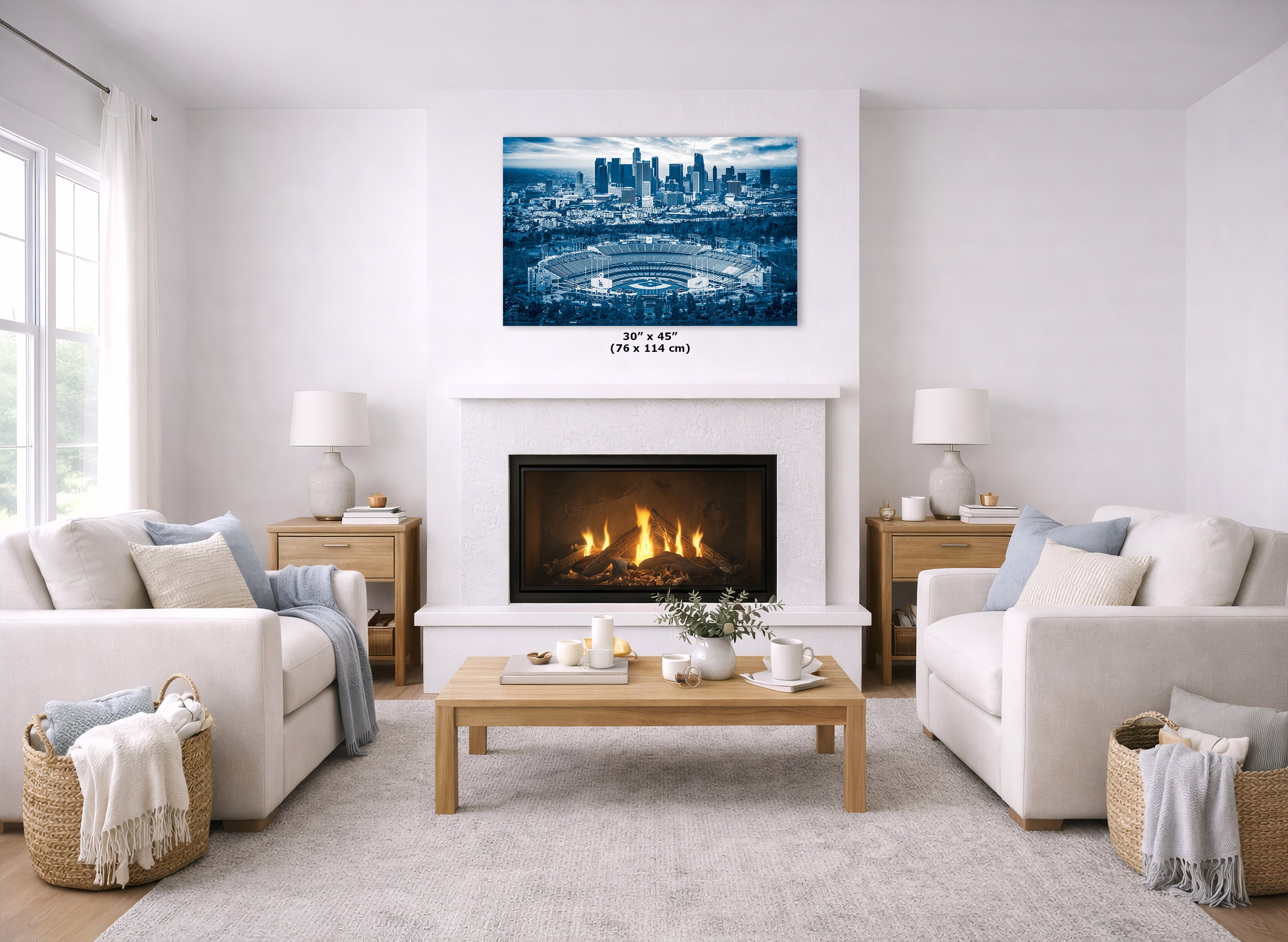 A square canvas wall art print depicting a blue-toned city skyline and stadium, displayed above a white fireplace in a bright, modern living room with two white sofas and a wooden coffee table.