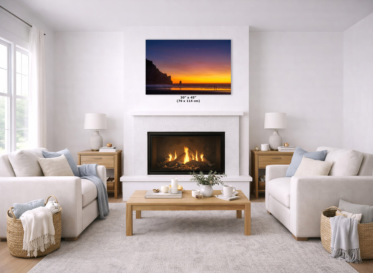 A wall art print showing a colorful sunset over water is displayed above a white fireplace in a bright, modern living room with two cream sofas, a wooden coffee table, and a light rug.