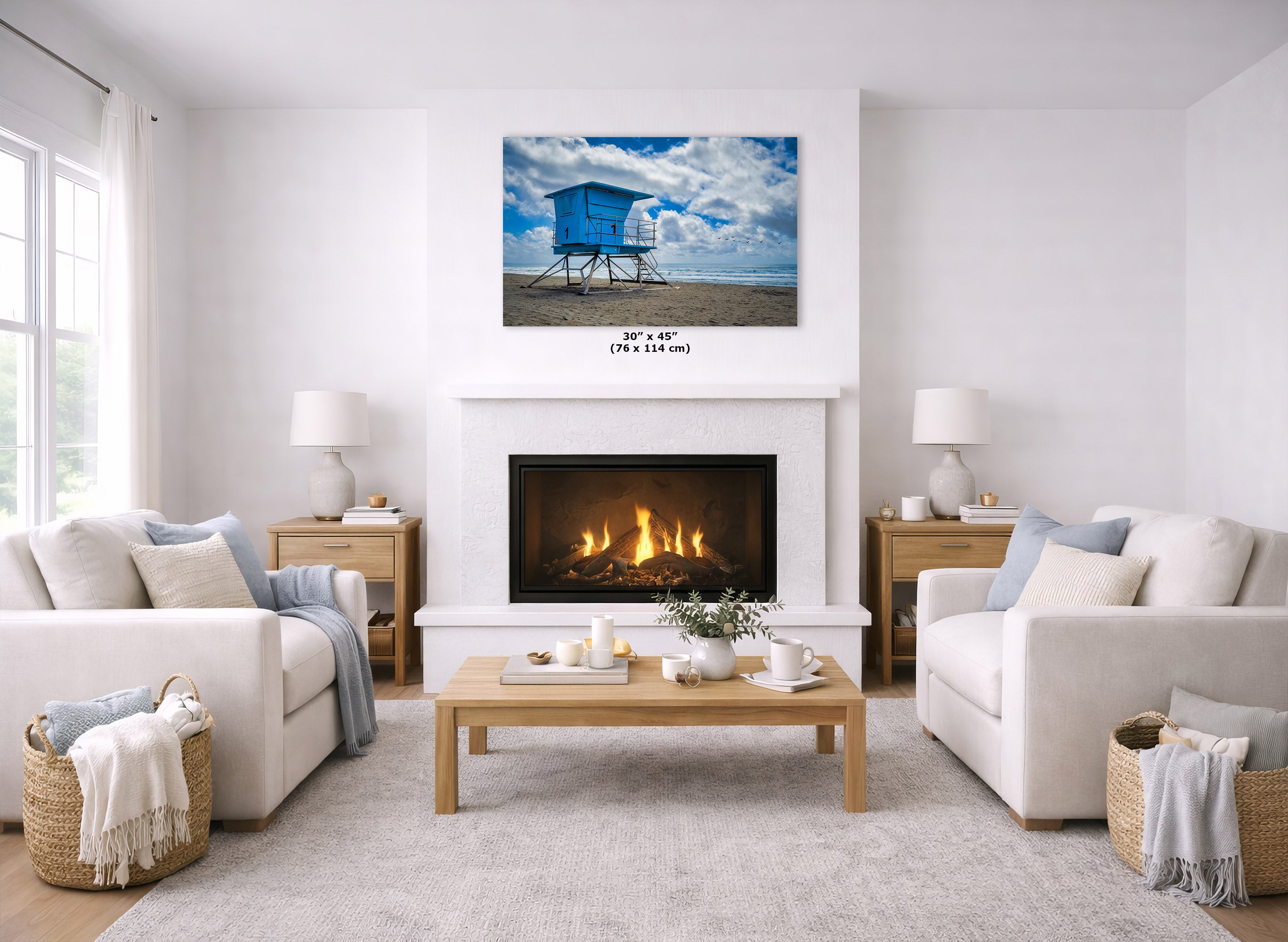 A wall art print showing a blue lifeguard hut on a beach under a cloudy sky, displayed above a fireplace in a bright, modern living room with white sofas, a wooden coffee table, and a light rug.