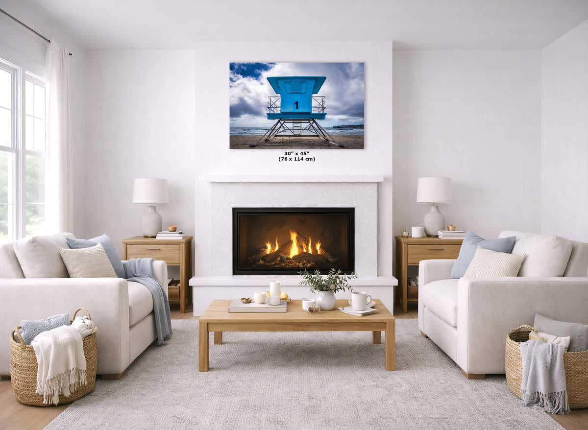 A wall art print showing a bright blue lifeguard tower over water, displayed above a fireplace in a bright, modern living room with white sofas, a wooden coffee table, and a light rug.