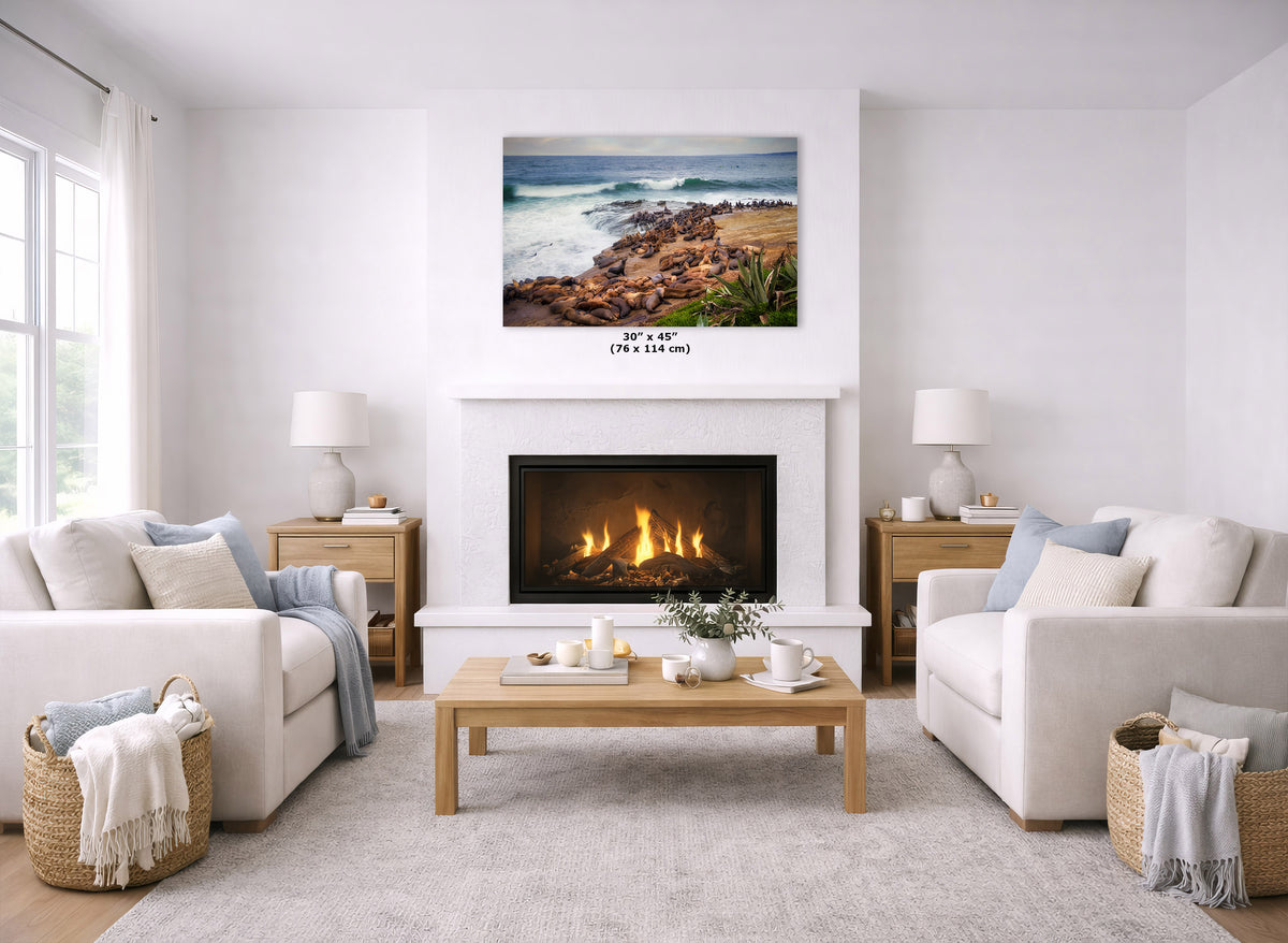 A coastal landscape print hangs above a white fireplace in a bright, modern living room with two cream sofas, a wooden coffee table, and woven baskets.