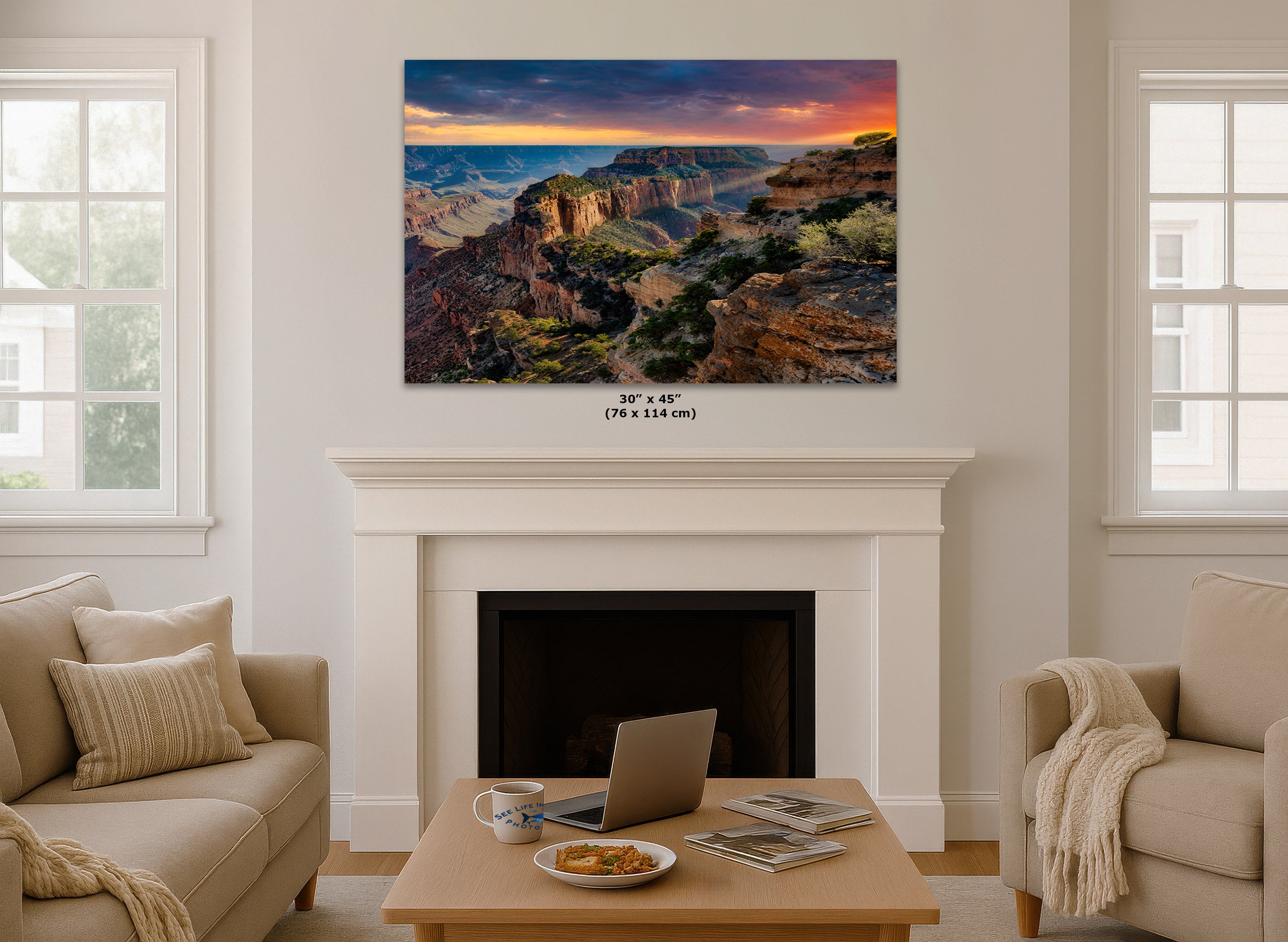 A large rectangular wall art print showing a Grand Canyon landscape at sunset hangs above a white fireplace in a bright living room. The print is the product being sold, but the room setting is visible, so this is best classified as Lifestyle rather than Scenery or Product/Packaging.