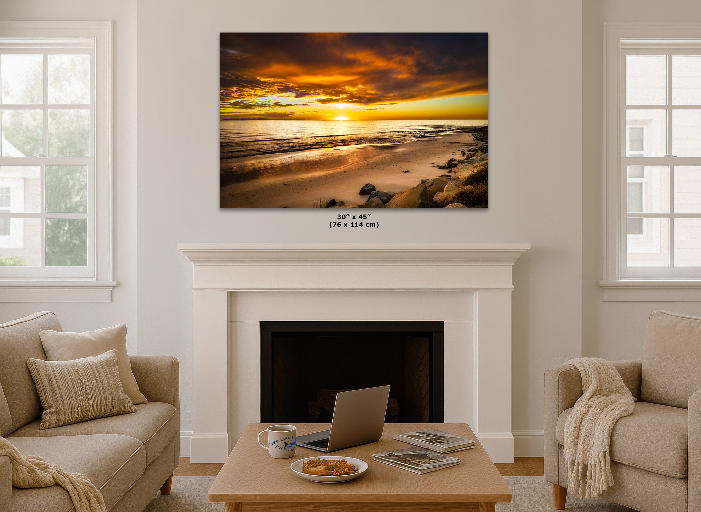 A wall art print showing a dramatic beach sunset with orange and yellow clouds, displayed above a white fireplace mantel in a bright living room. Small text below the print reads 30 x 45 in (76 x 114 cm).