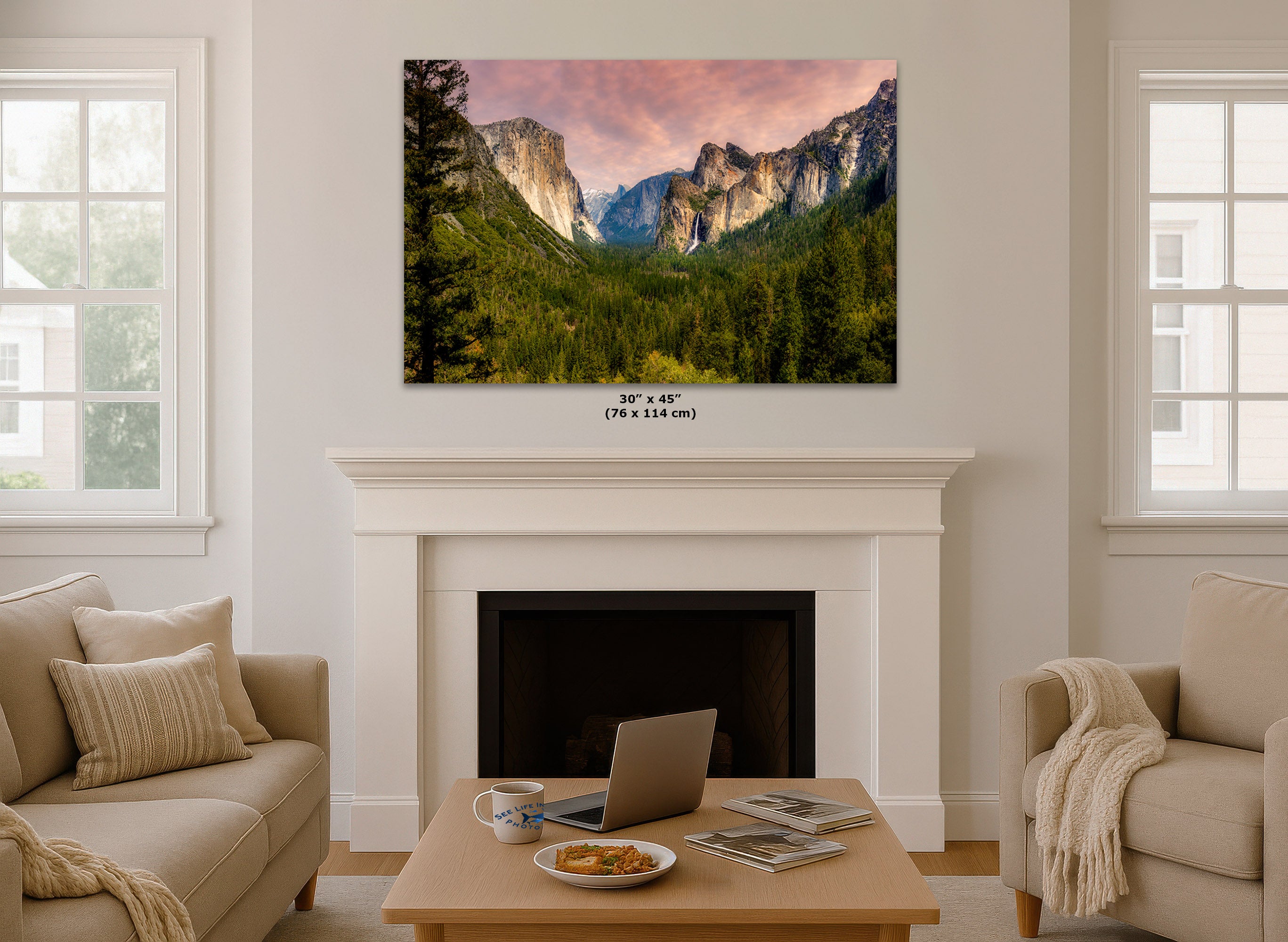A wall art print showing a mountain valley landscape with pink sky and green trees, displayed above a white fireplace in a bright living room. Small text below the print reads 30 x 45 in (76 x 114 cm)