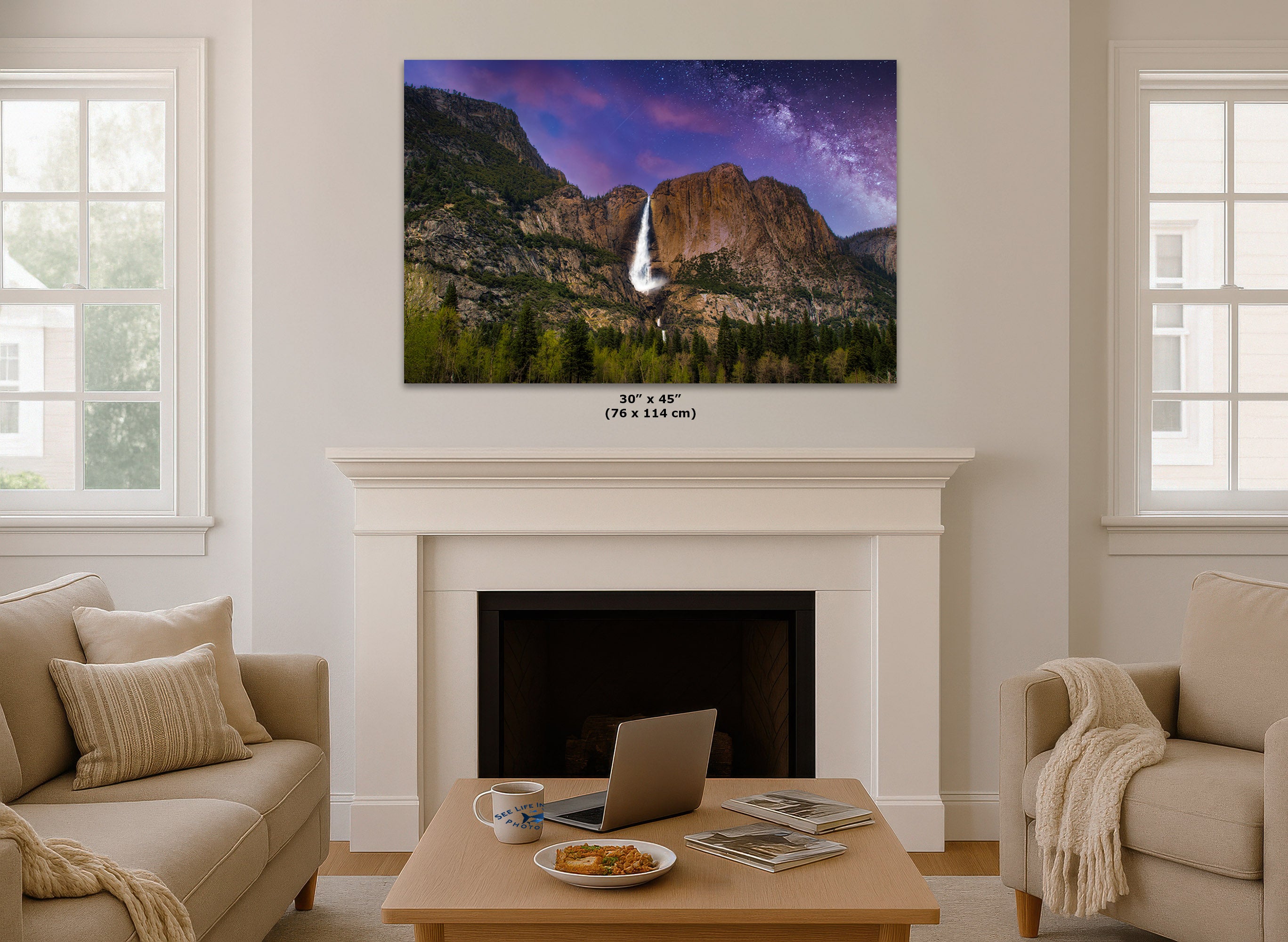 A wall art print showing a mountain landscape with a waterfall under a purple sky, displayed above a white fireplace in a bright living room. Small text below the print reads 30 in x 45 in (76 x 114 cm).