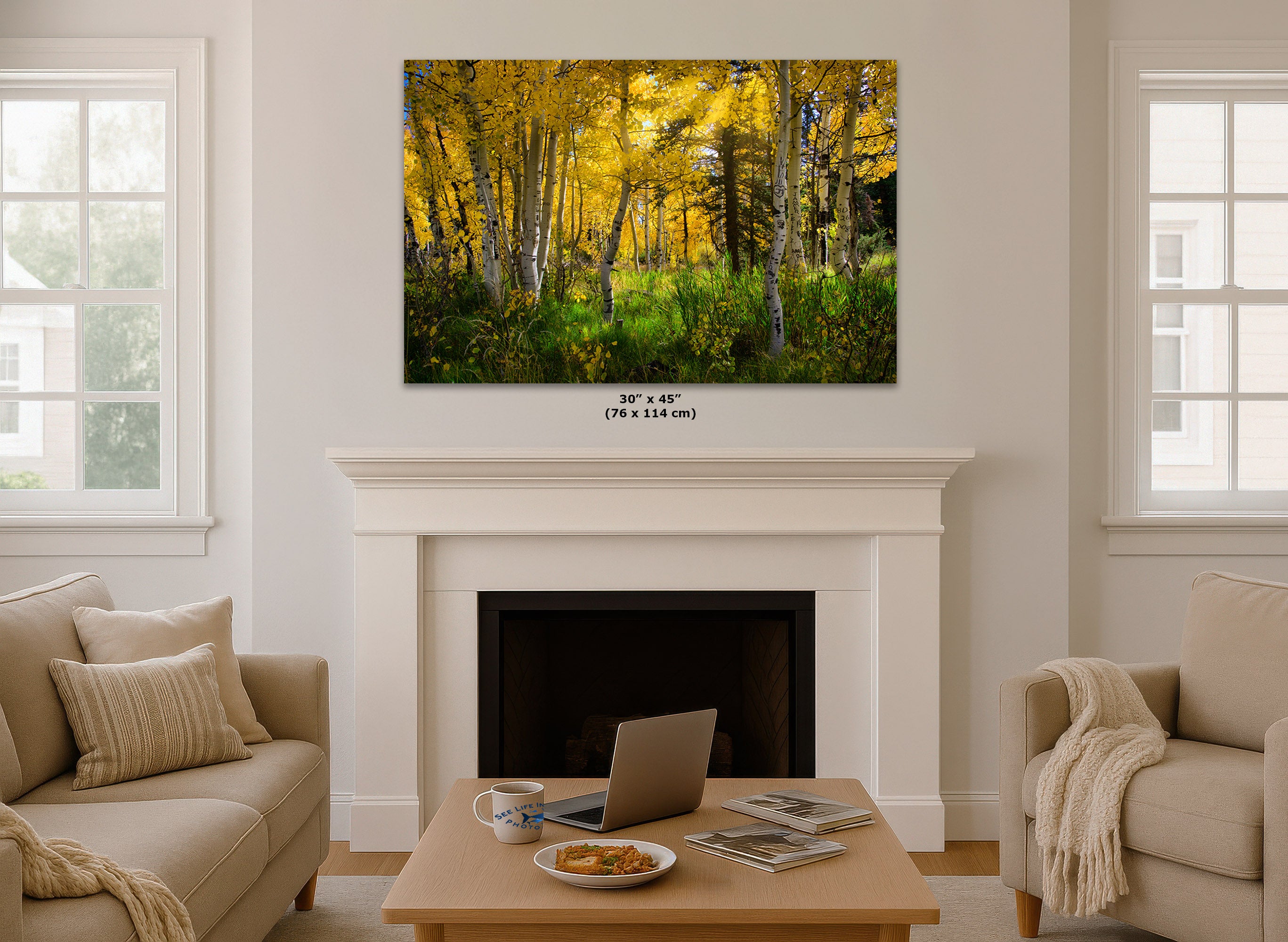 A wall art print showing a golden autumn forest hangs above a white fireplace mantel in a bright living room. The print depicts yellow and green trees with brown trunks.