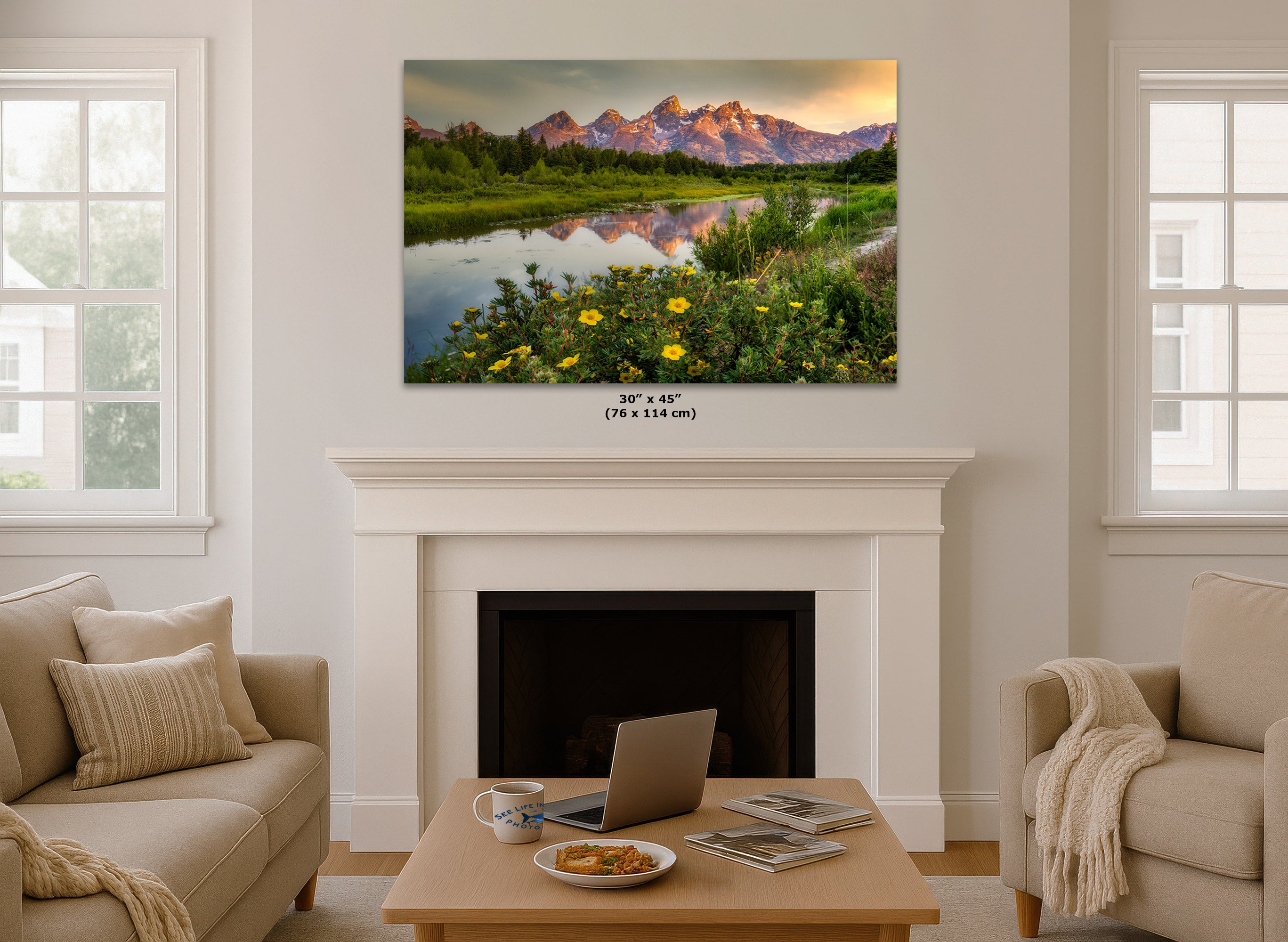 A wall art print showing a mountain landscape with a reflective lake and yellow wildflowers, displayed above a white fireplace in a bright living room. Small text below the print reads 30 x 45 (76 x 114 cm).