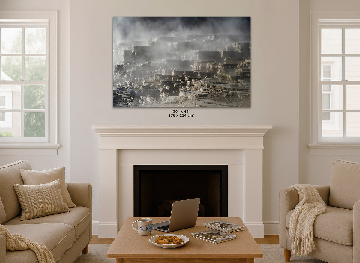 A large rectangular wall art print showing a smoky geothermal landscape in grayscale, displayed above a white fireplace mantel in a bright living room with beige sofas, a coffee table, and two windows.