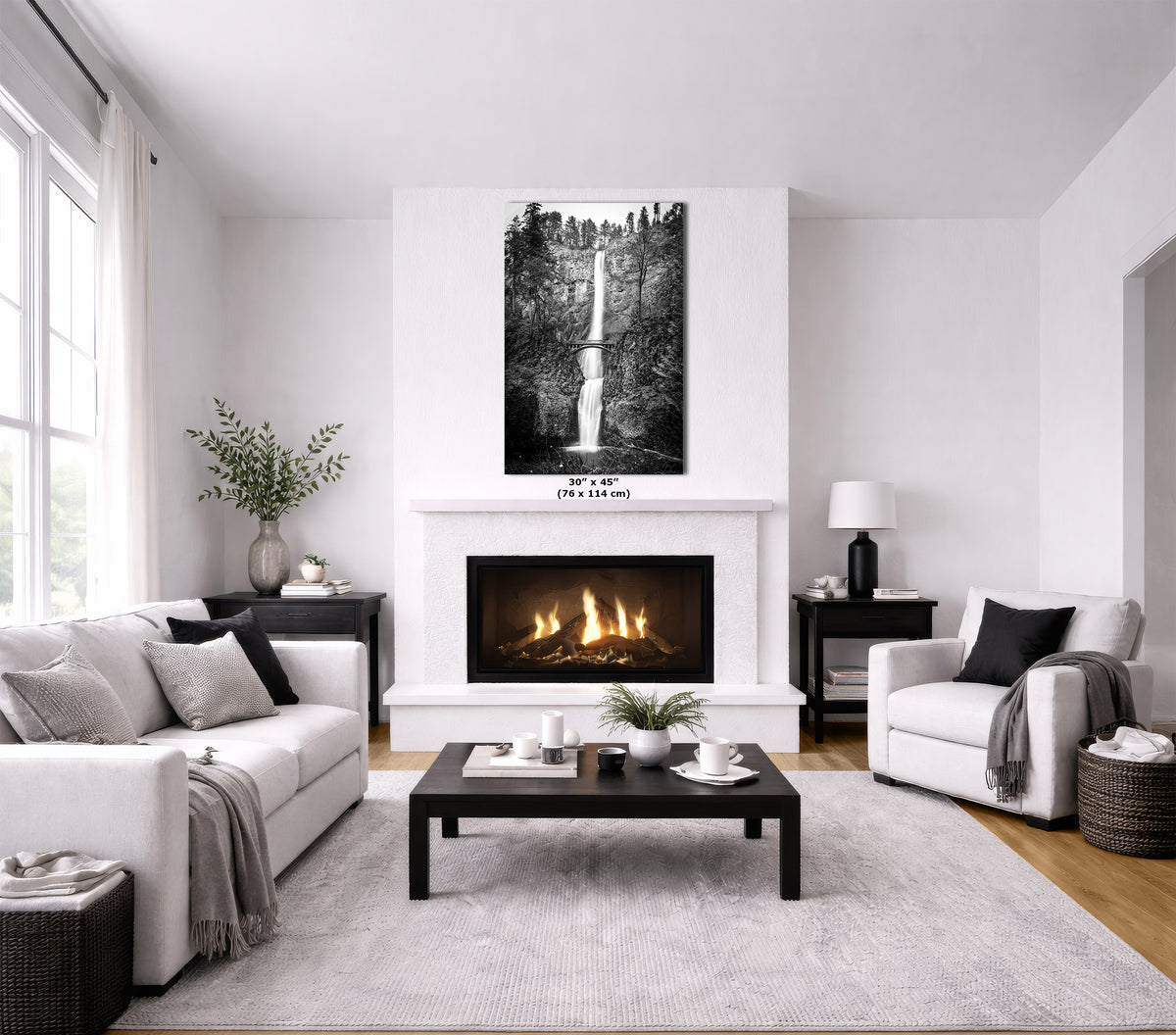 A modern living room with a white fireplace and wall, featuring a black-and-white waterfall wall art print above the fireplace. The room includes a white sofa, a white armchair, a dark wood coffee table, and a light gray rug.