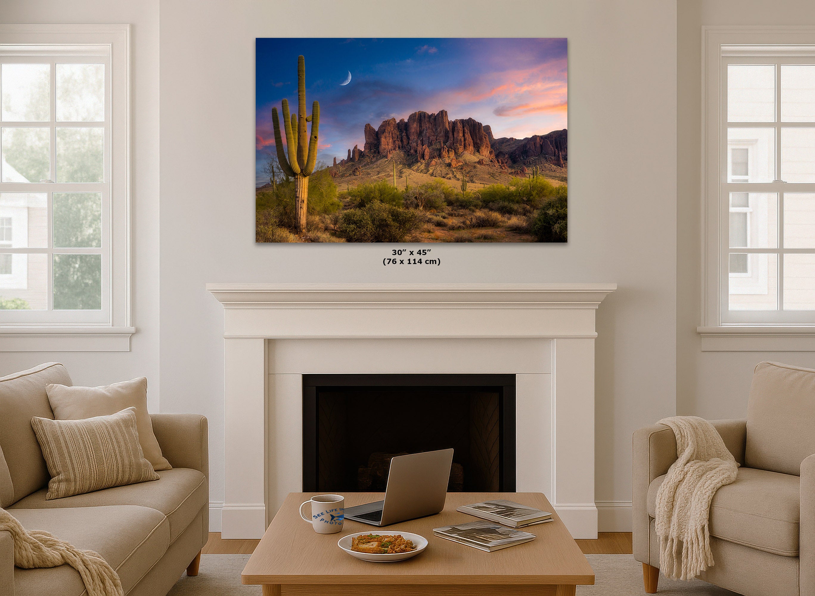 A desert landscape wall art print hangs above a white fireplace in a bright living room. The artwork shows a saguaro cactus, rocky mountains, and a colorful sunset sky with a crescent moon.