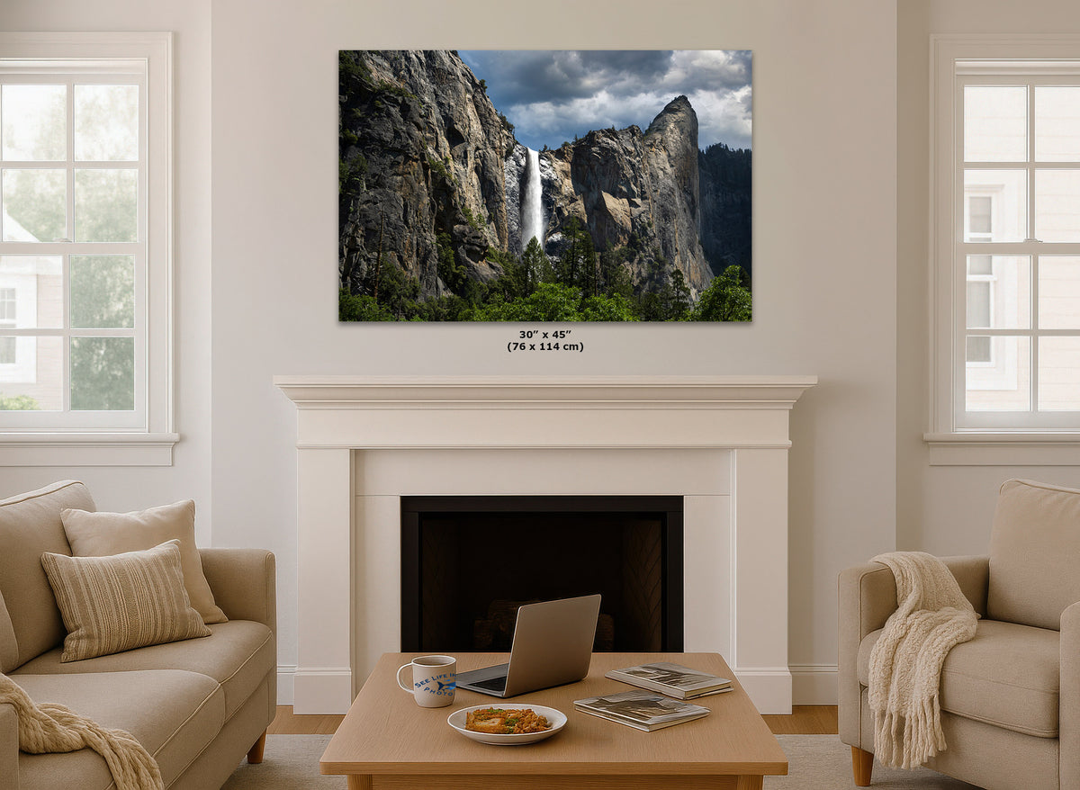 A wall art print of Yosemite Falls, shown hanging above a white fireplace mantel in a styled living room with beige sofas and a coffee table. The print includes a size label reading 30 x 45 in (76 x 114 cm).