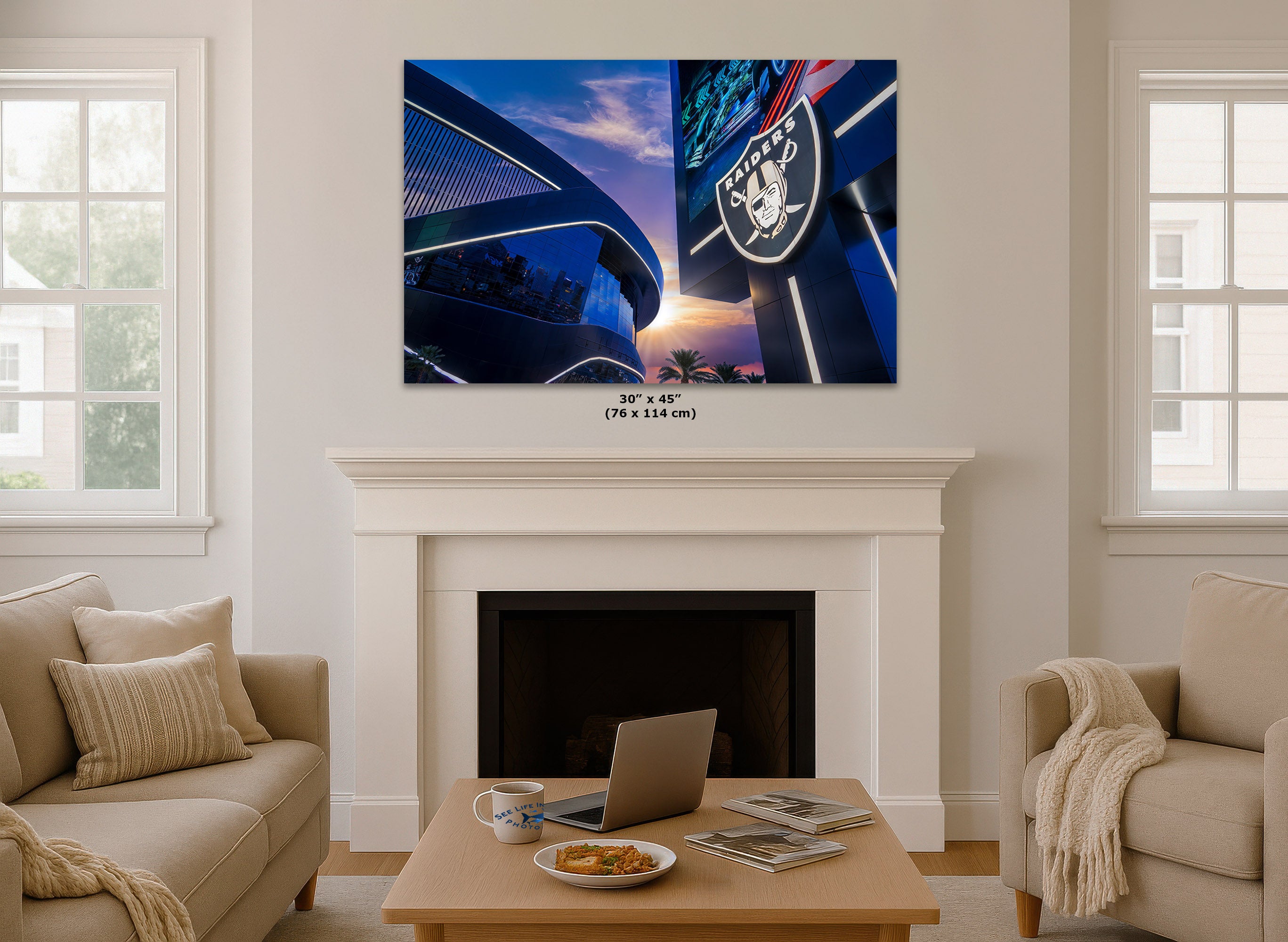 A framed wall art print showing a blue-toned stadium and Las Vegas sign is displayed above a white fireplace in a bright living room. The print includes the word 'RAIDERS' and a skull logo, and small text below reads 30" x 45" (76 x 114 cm).