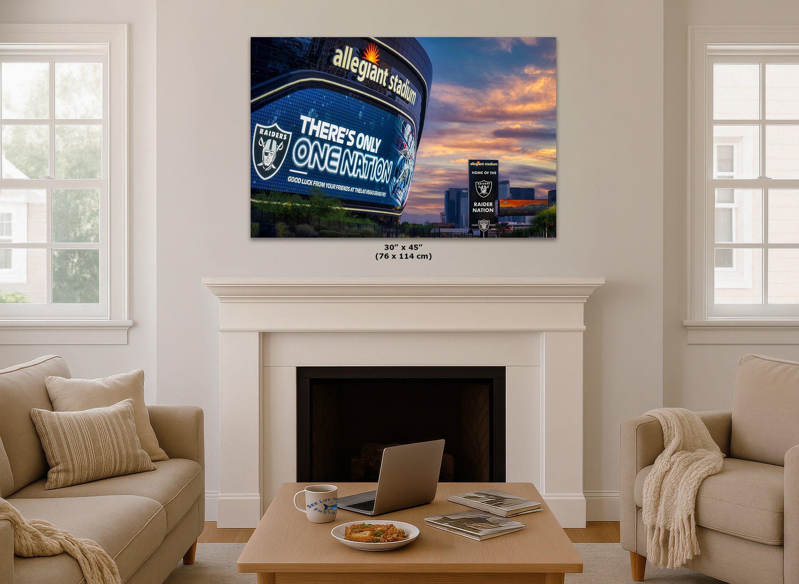 A 30x45 inch canvas print of the Las Vegas Raiders and Allegiant Stadium sunset is displayed on a white wall above a white fireplace mantel in a modern living room setting.