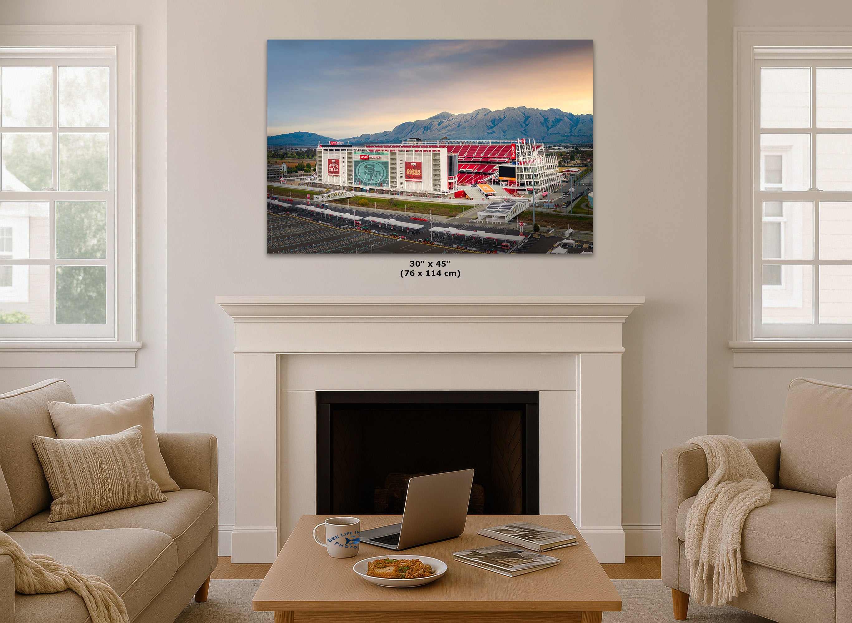 A wall art print showing a stadium and mountains is displayed above a white fireplace mantel in a bright living room. The print includes size text reading 30 x 45 in (76 x 114 cm).
