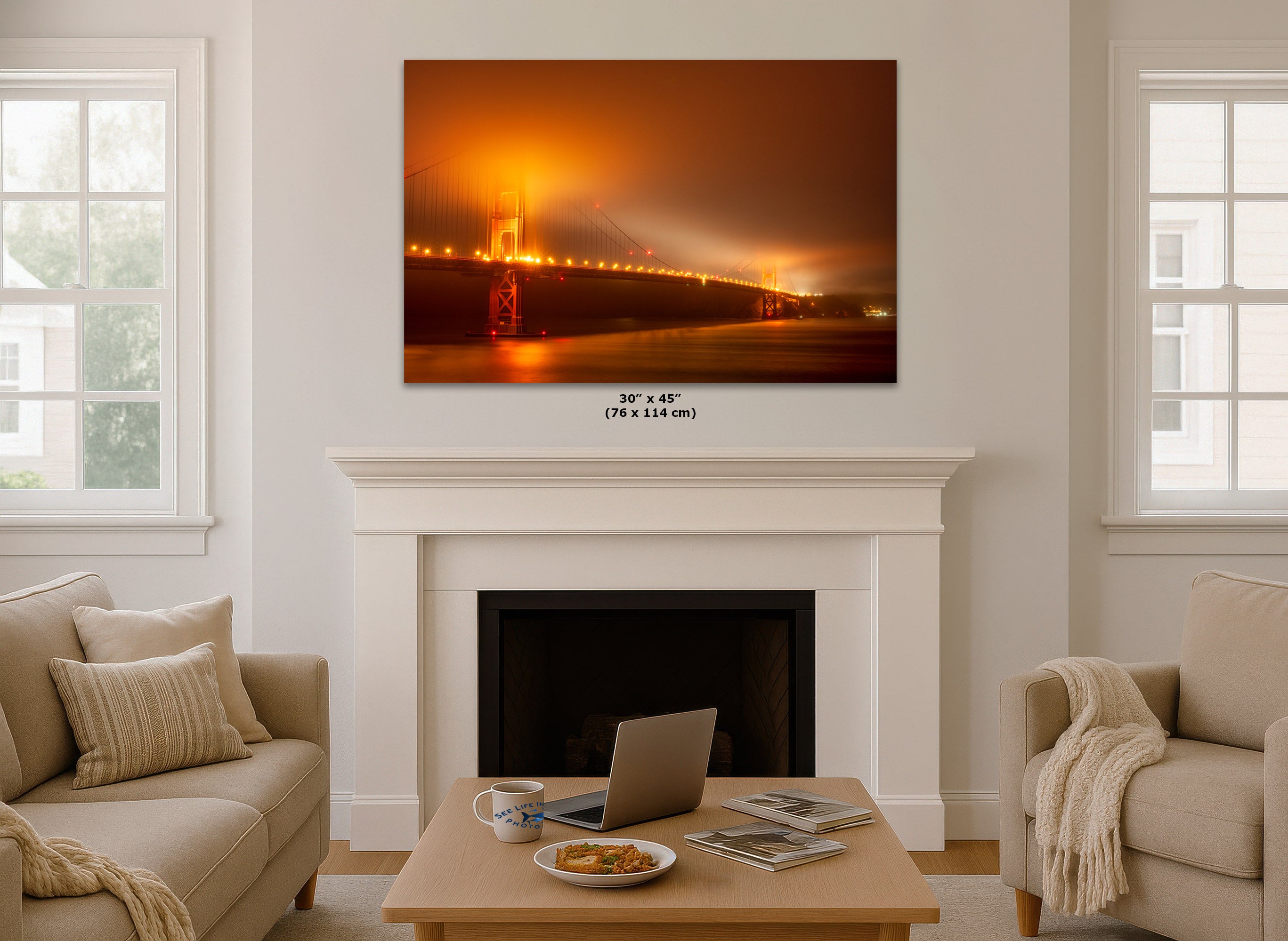 A limited edition wall art print showing a glowing orange Golden Gate Bridge at night hangs above a white fireplace in a bright living room. Small text below the print reads 30 x 45 (76 x 114 in).