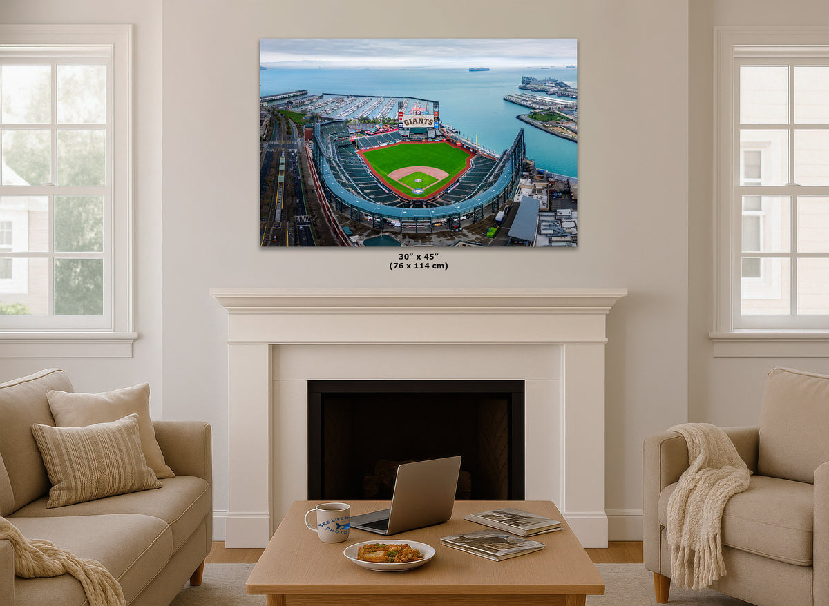A wall art print showing an aerial view of the Oracle Park baseball stadium by the water, displayed above a white fireplace mantel in a bright living room. The print includes size text below it reading 30 x 45 (76 x 114 in).