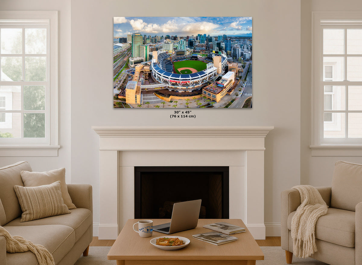 A 30" x 45" canvas print of a baseball stadium cityscape is displayed on a white fireplace mantel in a modern living room setting with beige sofas and a wooden coffee table.