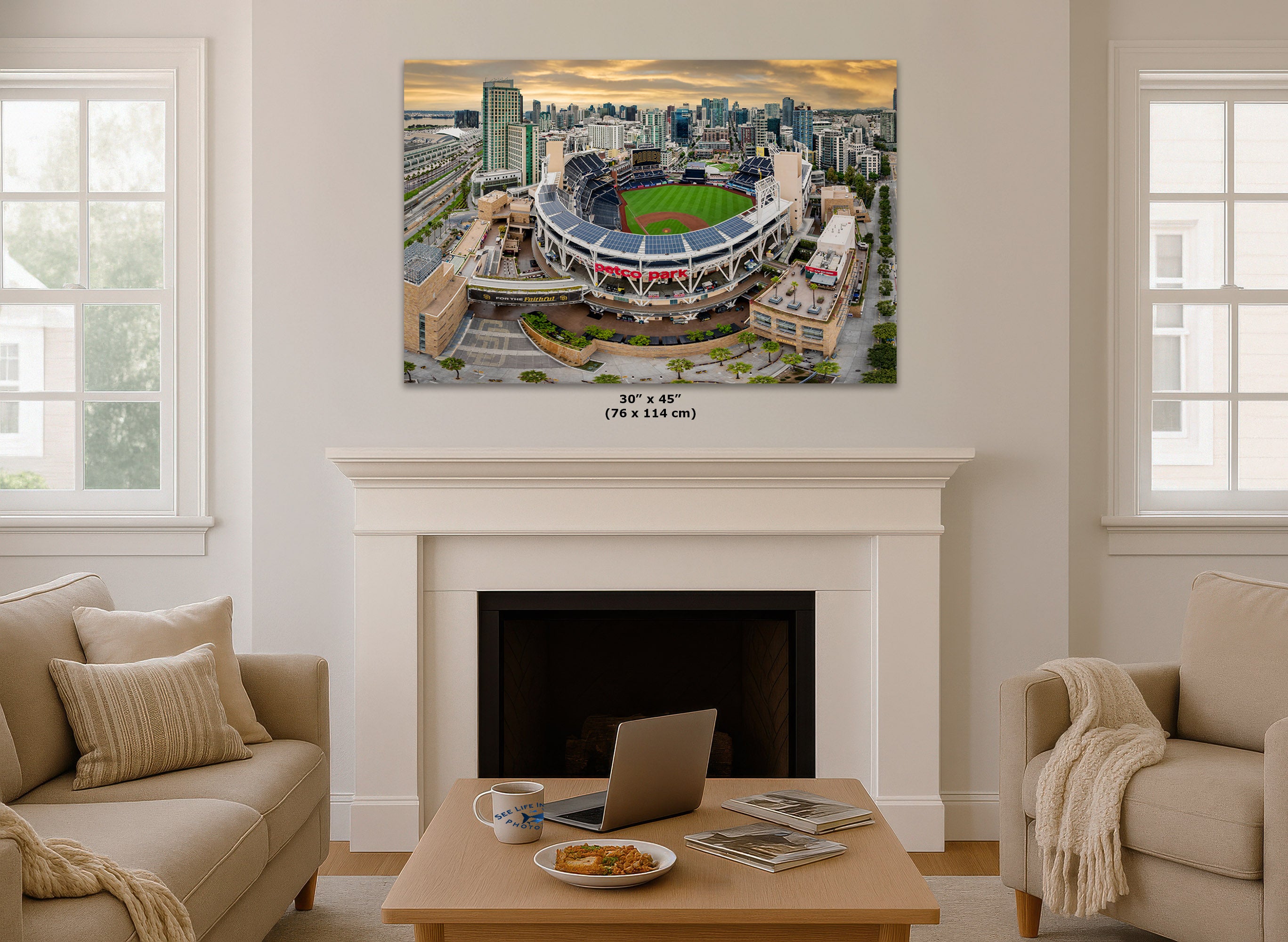 A large rectangular wall art print showing an aerial view of a stadium and city skyline, displayed above a white fireplace in a bright living room with beige sofas and a coffee table.