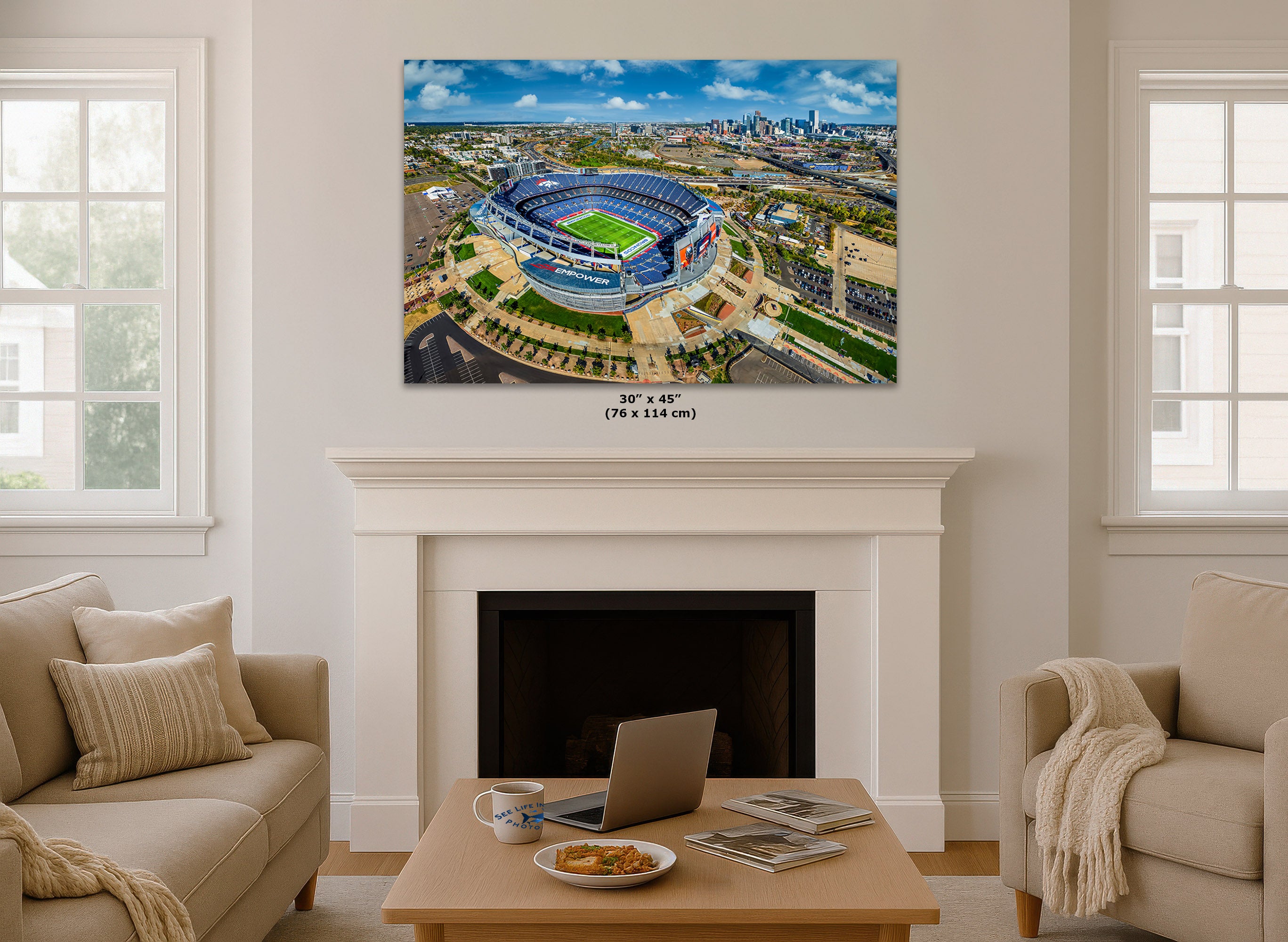 A large wall art print showing an aerial view of a stadium, displayed above a white fireplace mantel in a bright living room with beige sofas and a coffee table.