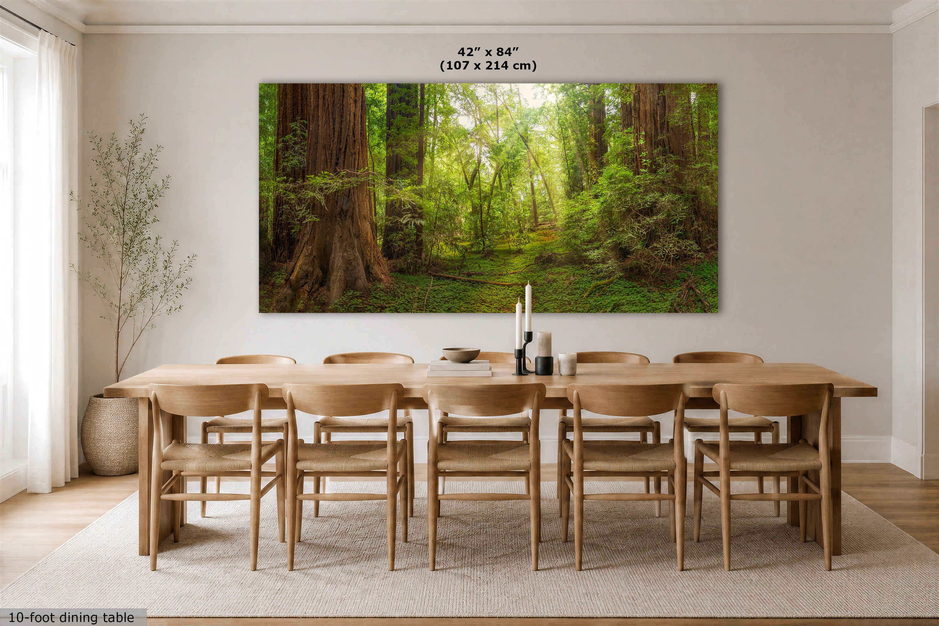 A large rectangular wall art print showing a sunlit forest path with tall trees and lush greenery, displayed above a wooden dining table and chairs in a bright dining room. Text at the top reads 42 x 84 inches (107 x 214 cm), and a small caption at bottom left says 10-foot dining table.