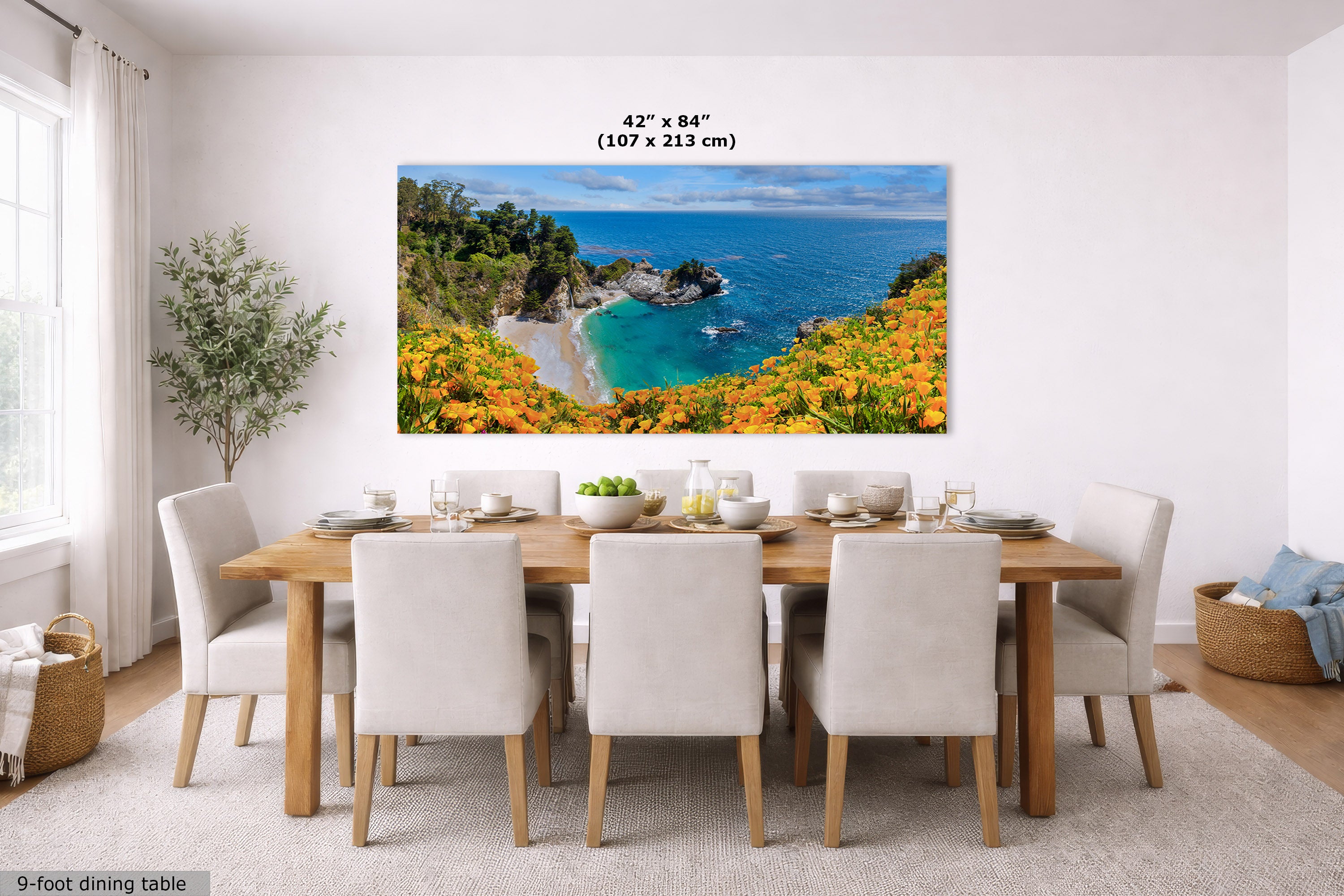 A large rectangular wall art print showing a coastal beach and ocean landscape with bright orange flowers in the foreground, displayed above a dining table in a bright modern room. Text above the print reads 42" x 84" (107 x 213 cm).