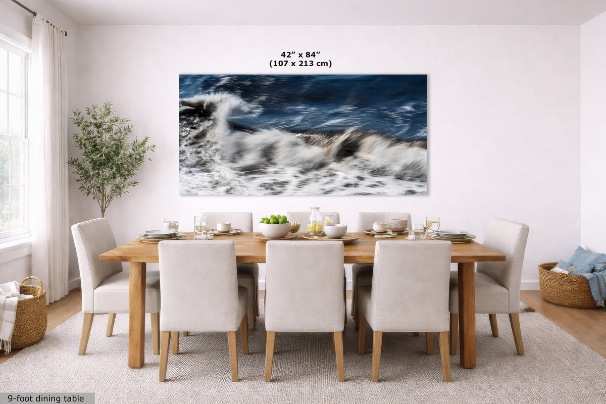 A large rectangular wall art print showing a dramatic ocean wave scene in deep blue and white tones, displayed above a wooden dining table with upholstered chairs in a bright dining room. Text at the top of the print reads 42" x 84" (107 x 213 cm), and a small label in the lower left says 9-foot dining table.