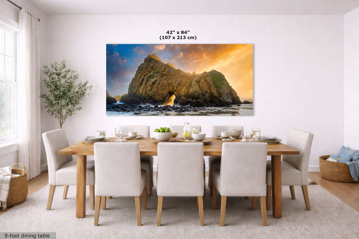 A large rectangular wall art print showing a rocky coastal cliff at sunset hangs above a dining table in a bright modern room. 