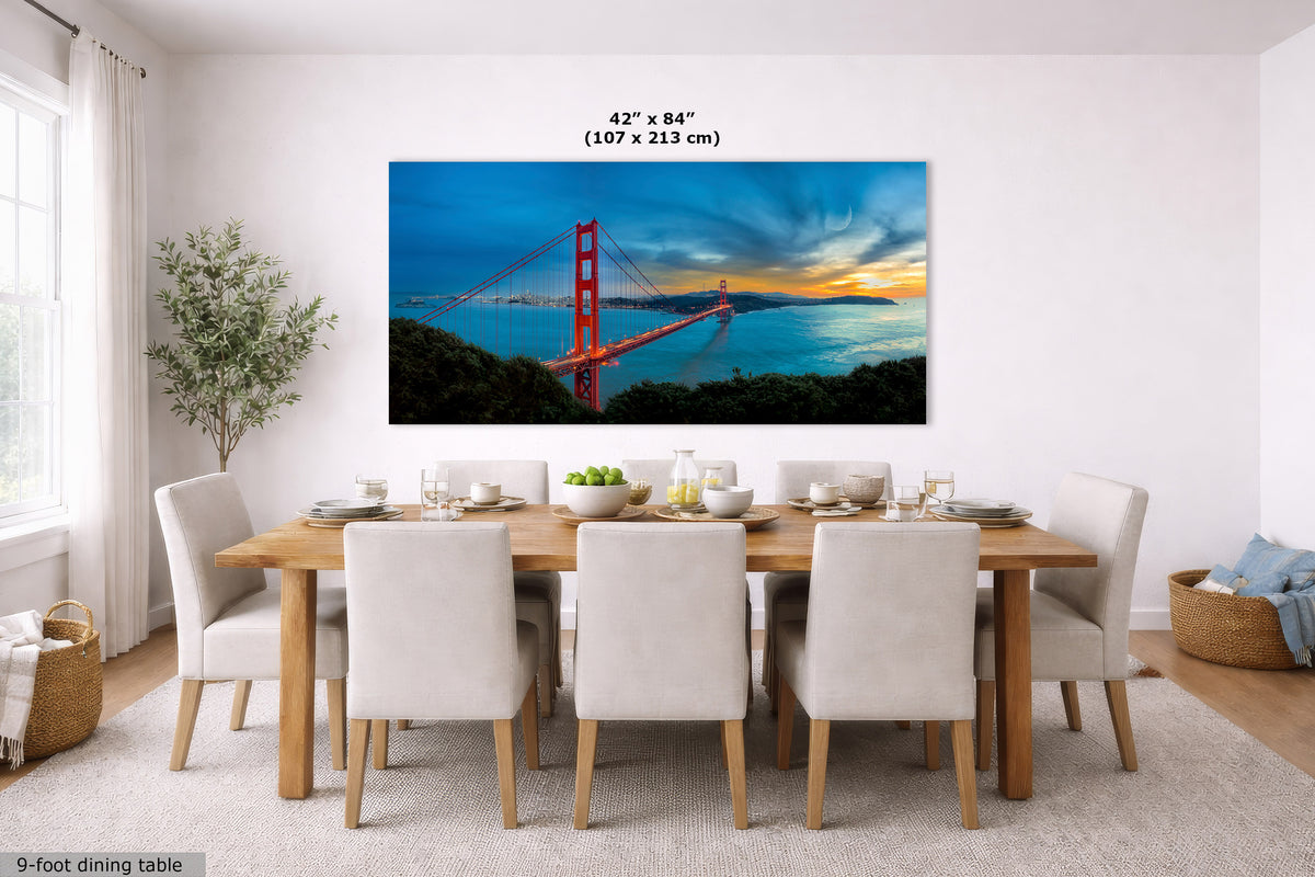 A large rectangular wall art print showing the Golden Gate Bridge over water at sunset, displayed above a dining table in a bright modern room. The print includes size text reading 42 x 84 inches (107 x 213 cm).