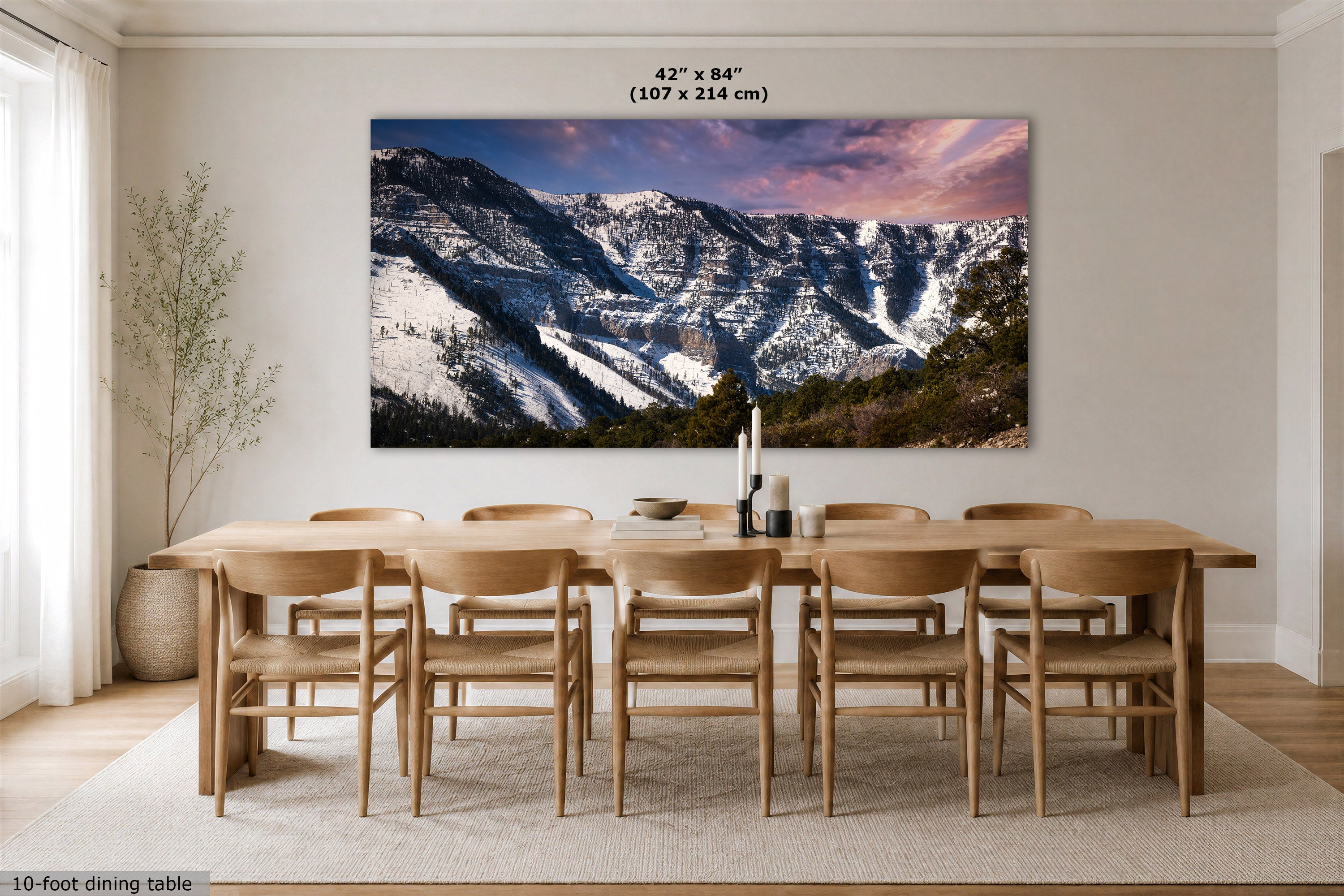 A large rectangular wall art print showing snowy mountain slopes under a pink and purple sky, displayed above a wooden dining table and chairs in a bright dining room. The print includes size text at the top: 42 x 84 inches (107 x 214 cm).