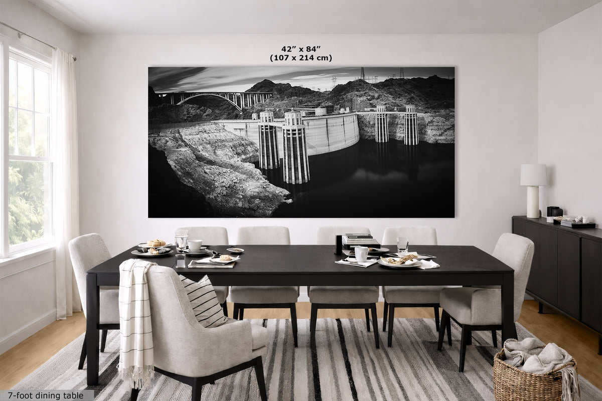 A large black and white wall art print depicting a dam and bridge landscape, displayed in a modern dining room setting above a dark wood dining table. The image includes a size dimension overlay at the top of the print.