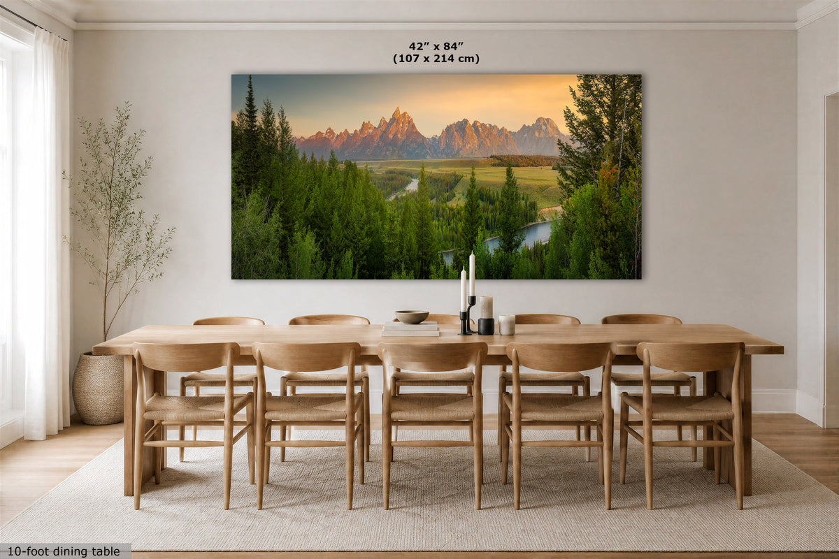 A large rectangular wall art print showing a mountain landscape with green trees and a river, displayed above a wooden dining table and chairs in a bright dining room. The print includes size text at the top and a small label at the bottom left.