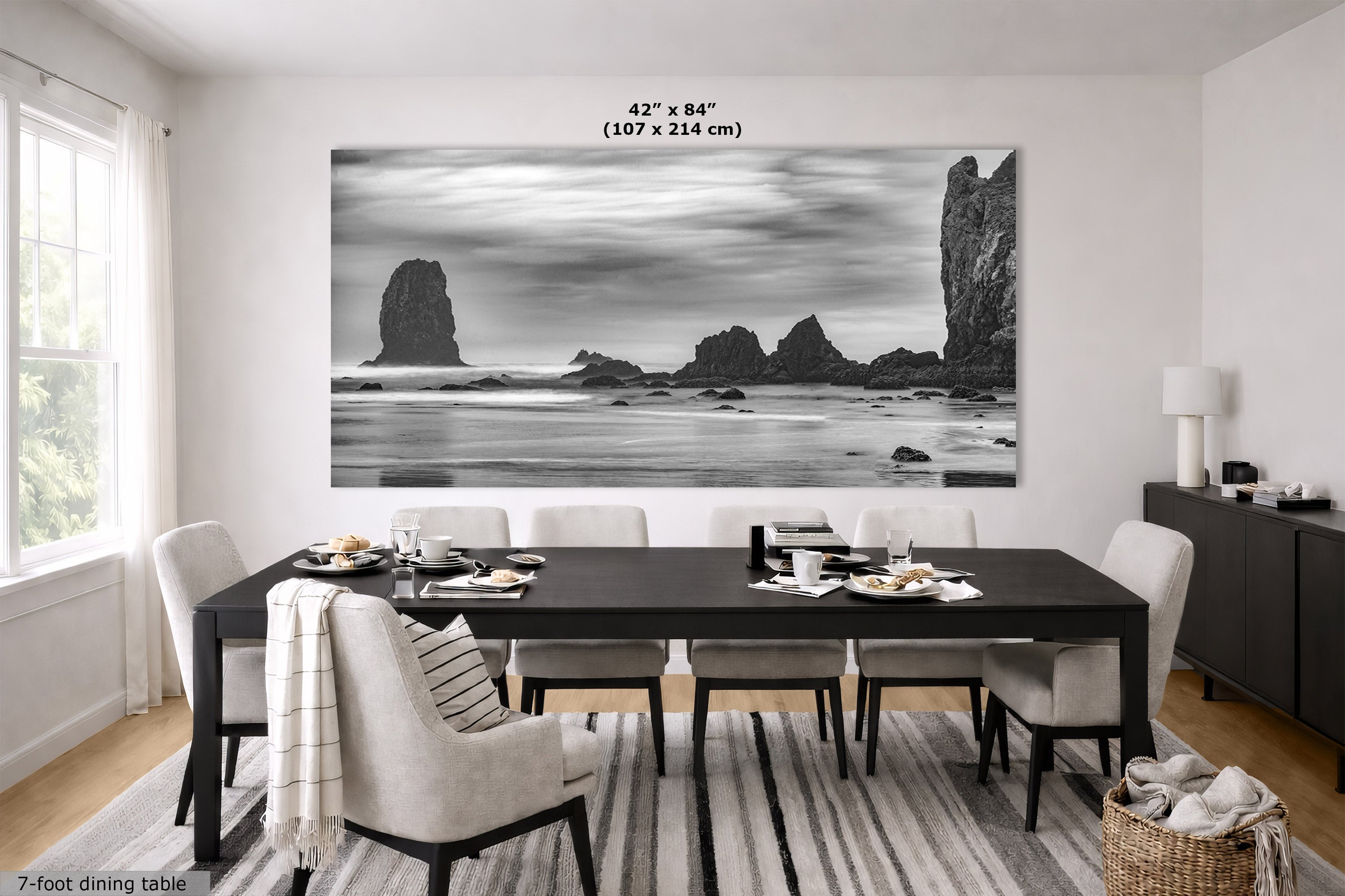 A large black and white coastal landscape wall art print hangs above a dark dining table with chairs in a bright modern dining room. The print depicts sea stacks and rocky cliffs over water.