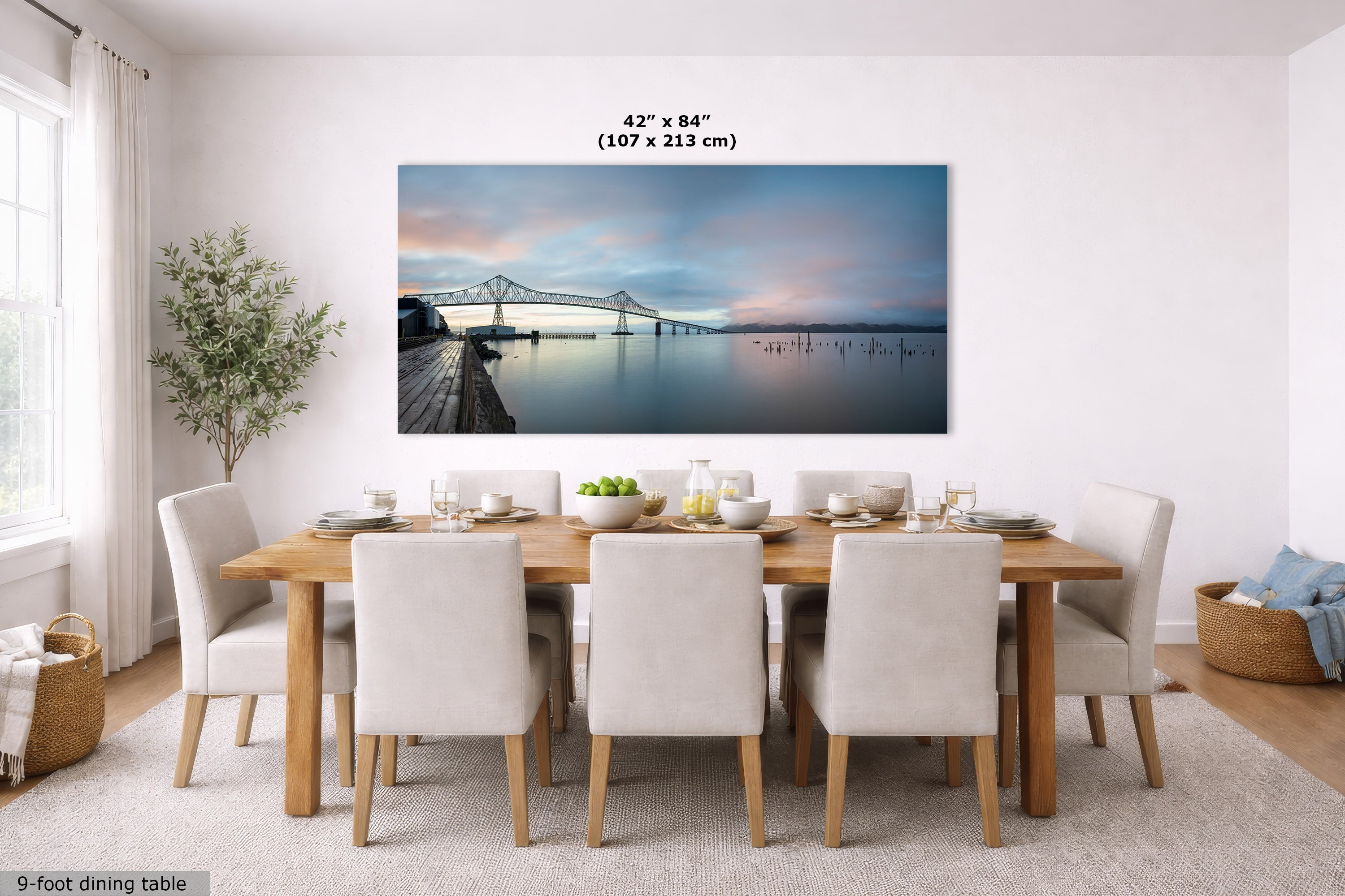 A large rectangular wall art print showing a waterfront bridge at dusk, displayed above a dining table in a bright modern dining room. The print includes size text reading 42 x 84 inches (107 x 213 cm).