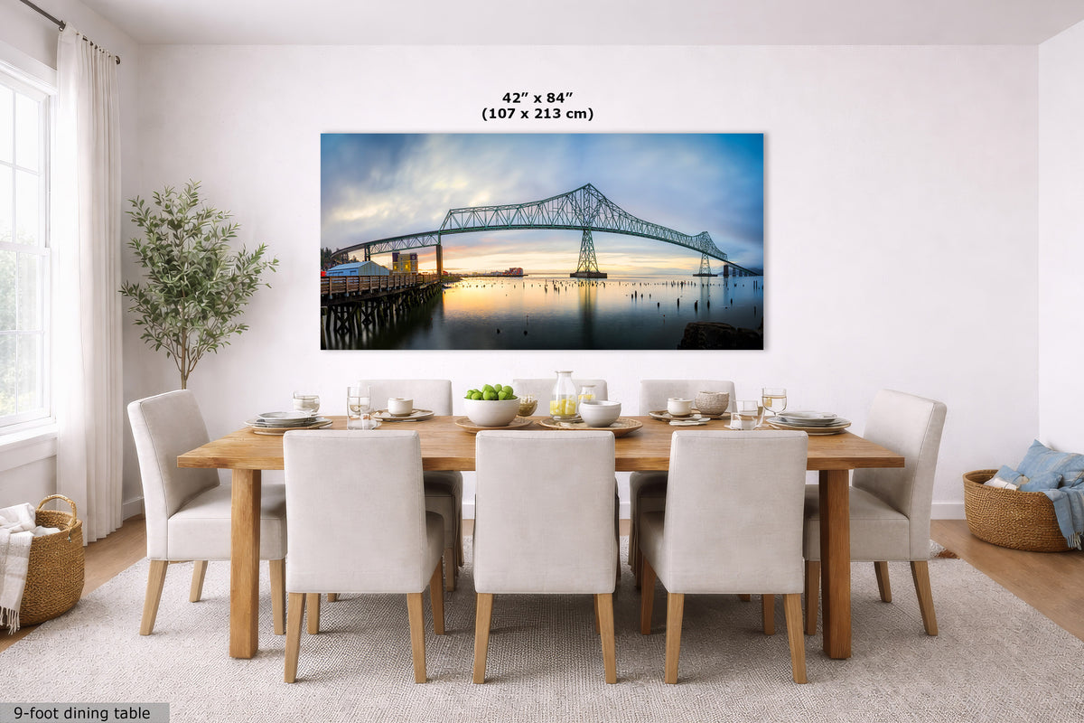 A large rectangular wall art print showing a bridge over water at dusk, displayed above a dining table in a bright modern room. The print includes size text reading 42 x 84 inches (107 x 213 cm).