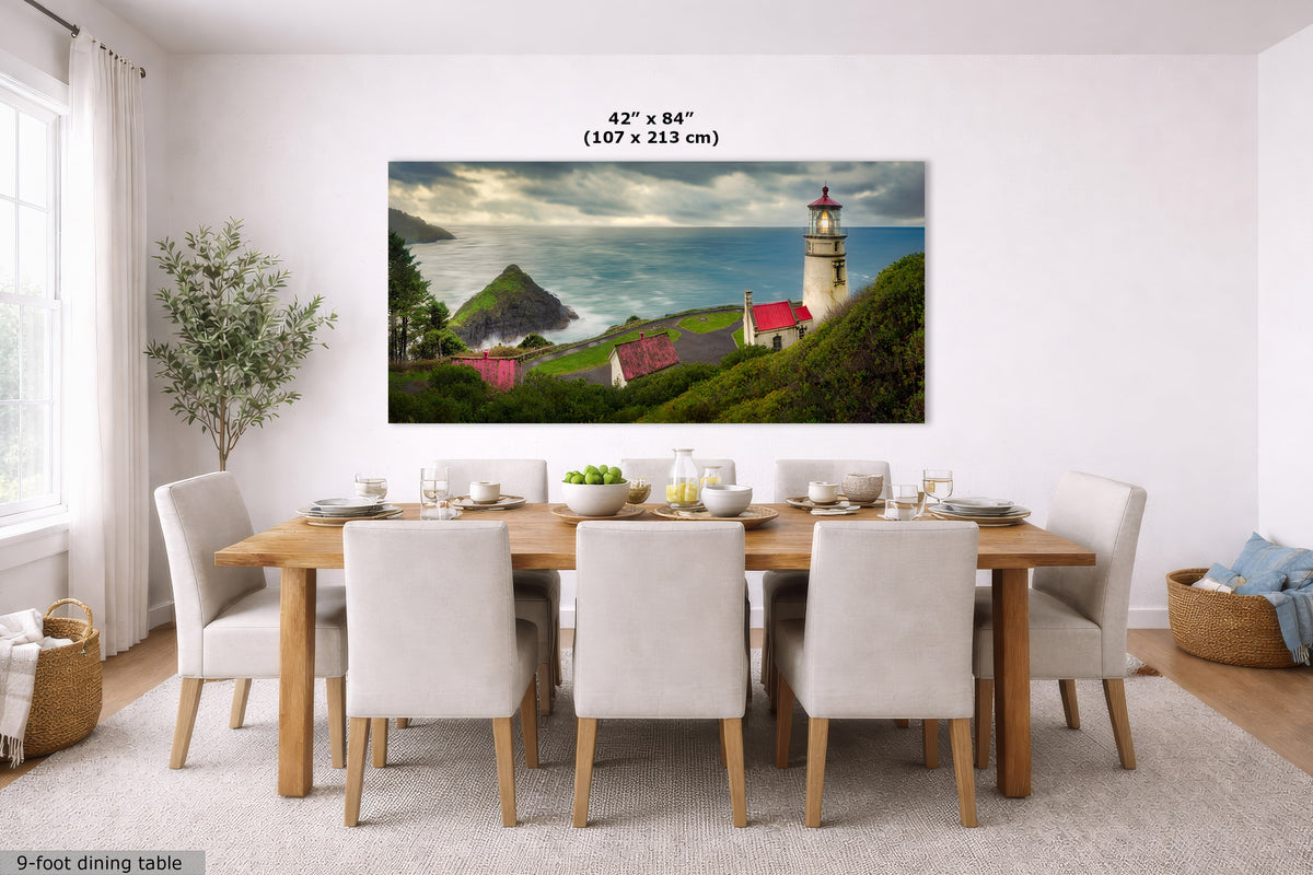 A large rectangular wall art print showing a coastal lighthouse scene with green hills, blue water, and a cloudy sky, displayed above a dining table in a bright modern room. Text at the top of the print reads 42" x 84" (107 x 213 cm).