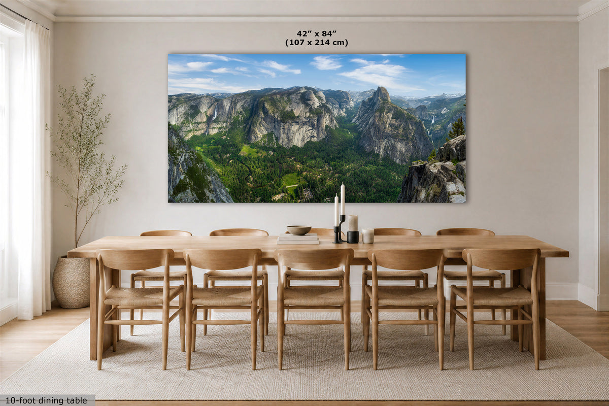 A large rectangular wall art print showing a mountain valley landscape with green forests and rocky peaks, displayed above a wooden dining table and chairs in a bright dining room. Text at the top reads 42" x 84" (107 x 214 cm), and text at the bottom left says 10-foot dining table.