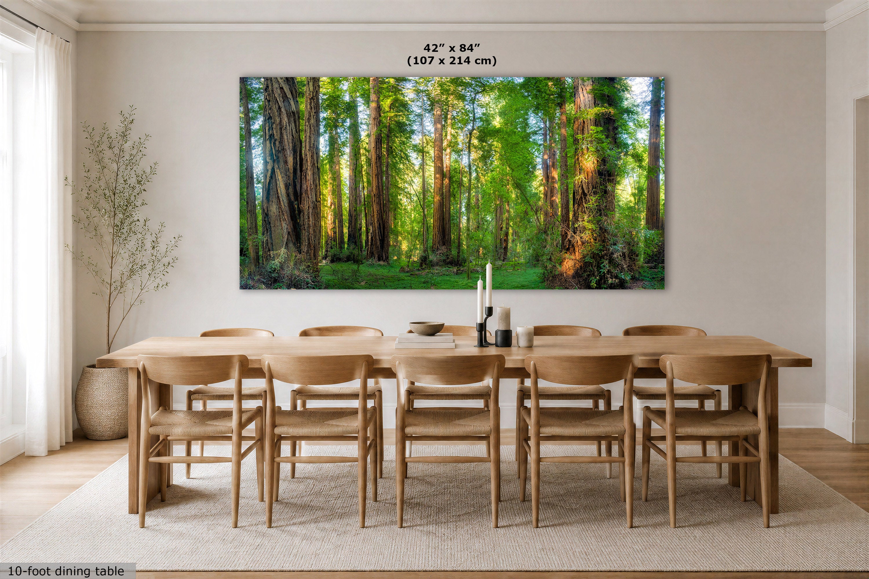 A large rectangular wall art print showing a sunlit forest of tall redwood trees, displayed above a wooden dining table and chairs in a bright dining room. Small text at the top reads 42 x 84" (107 x 214 cm), and text at the bottom left says 10-foot dining table.