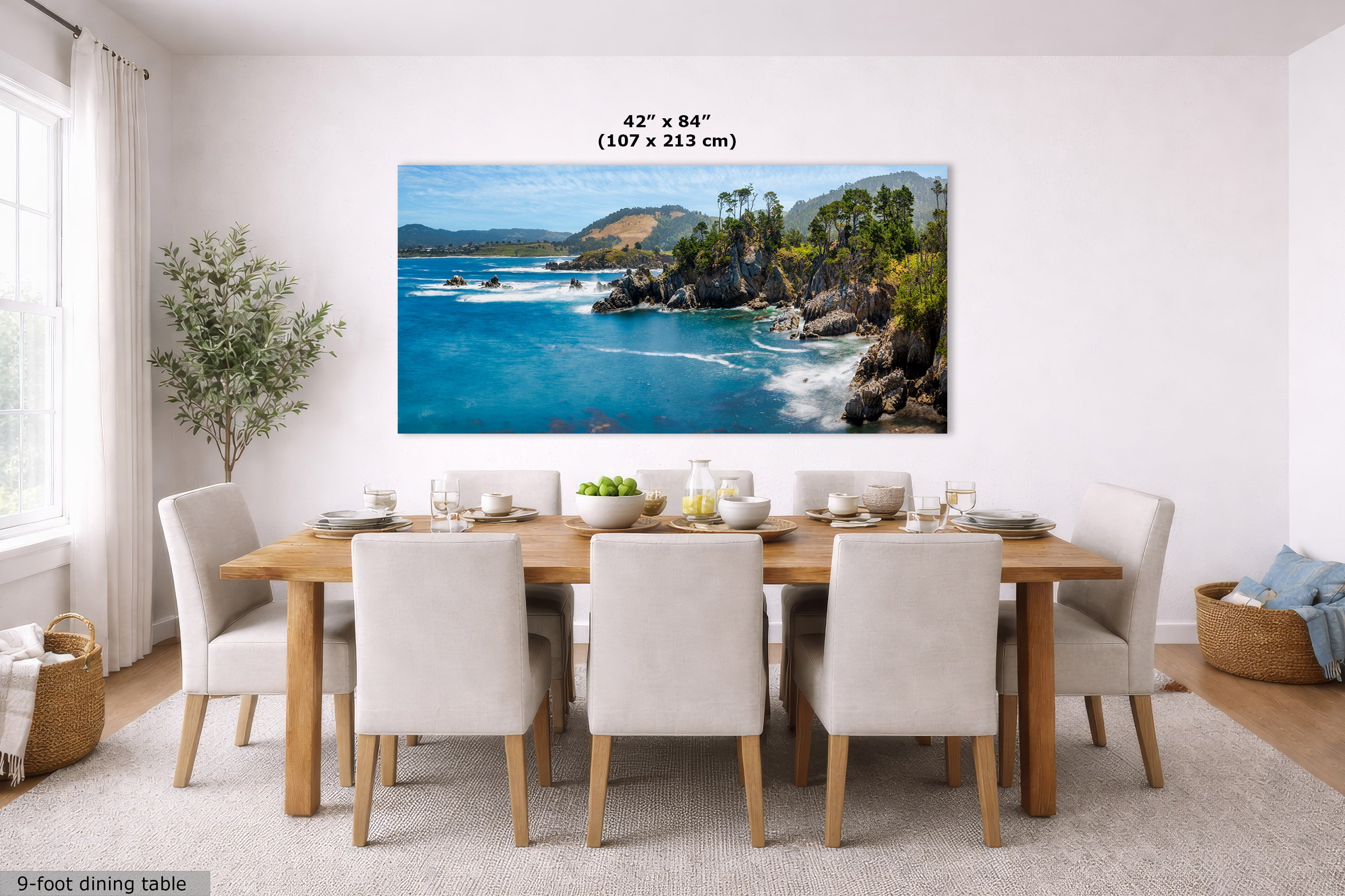 A large rectangular wall art print showing a coastal ocean scene with blue water, rocky cliffs, and green hills, displayed above a dining table in a bright modern room. Text above the print reads 42 x 84 inches (107 x 213 cm).