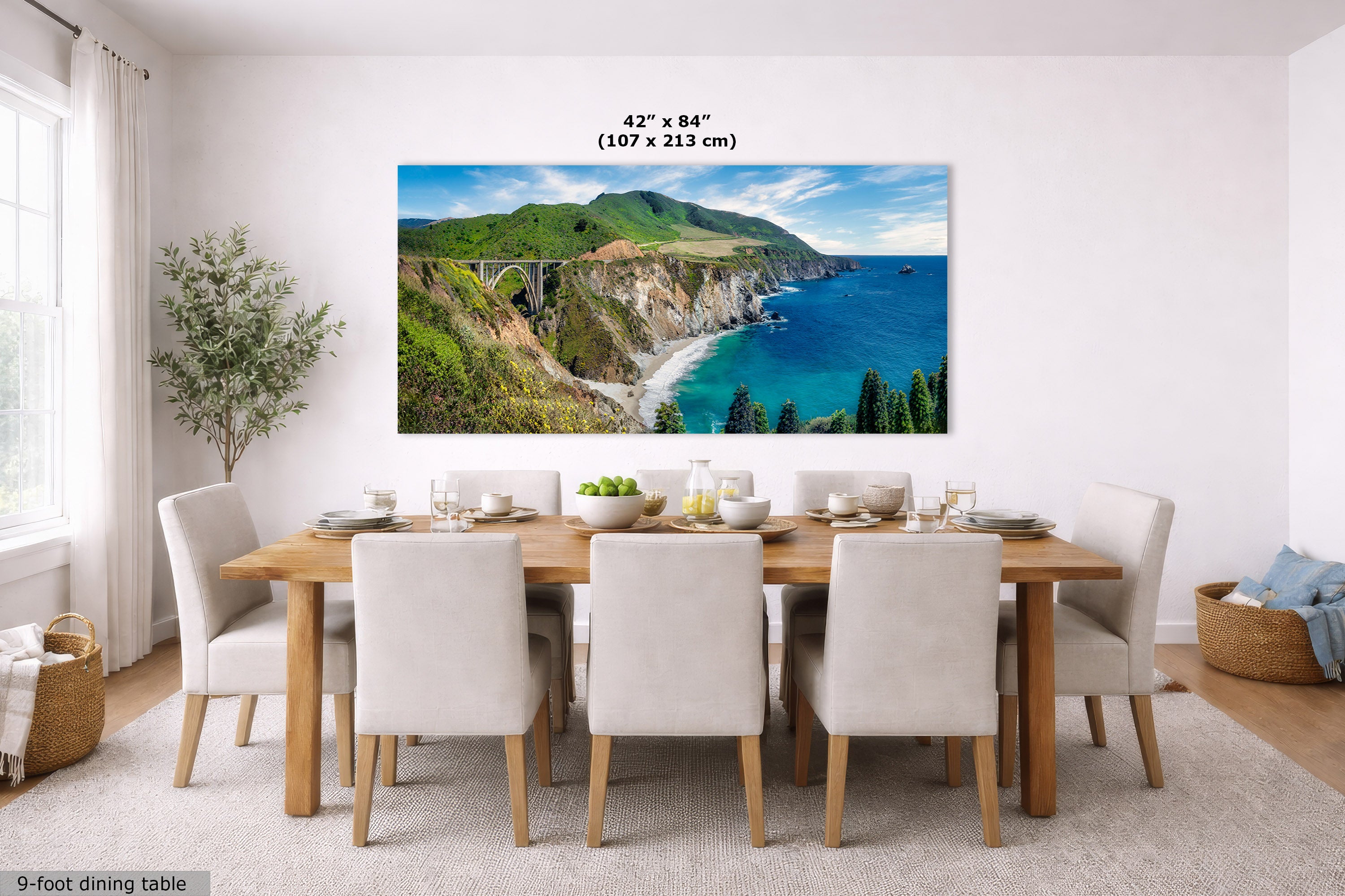 A large wall art print showing a coastal cliff and turquoise bay landscape, displayed above a dining table in a bright modern room. The print includes size text reading 42 x 84 inches (107 x 213 cm).