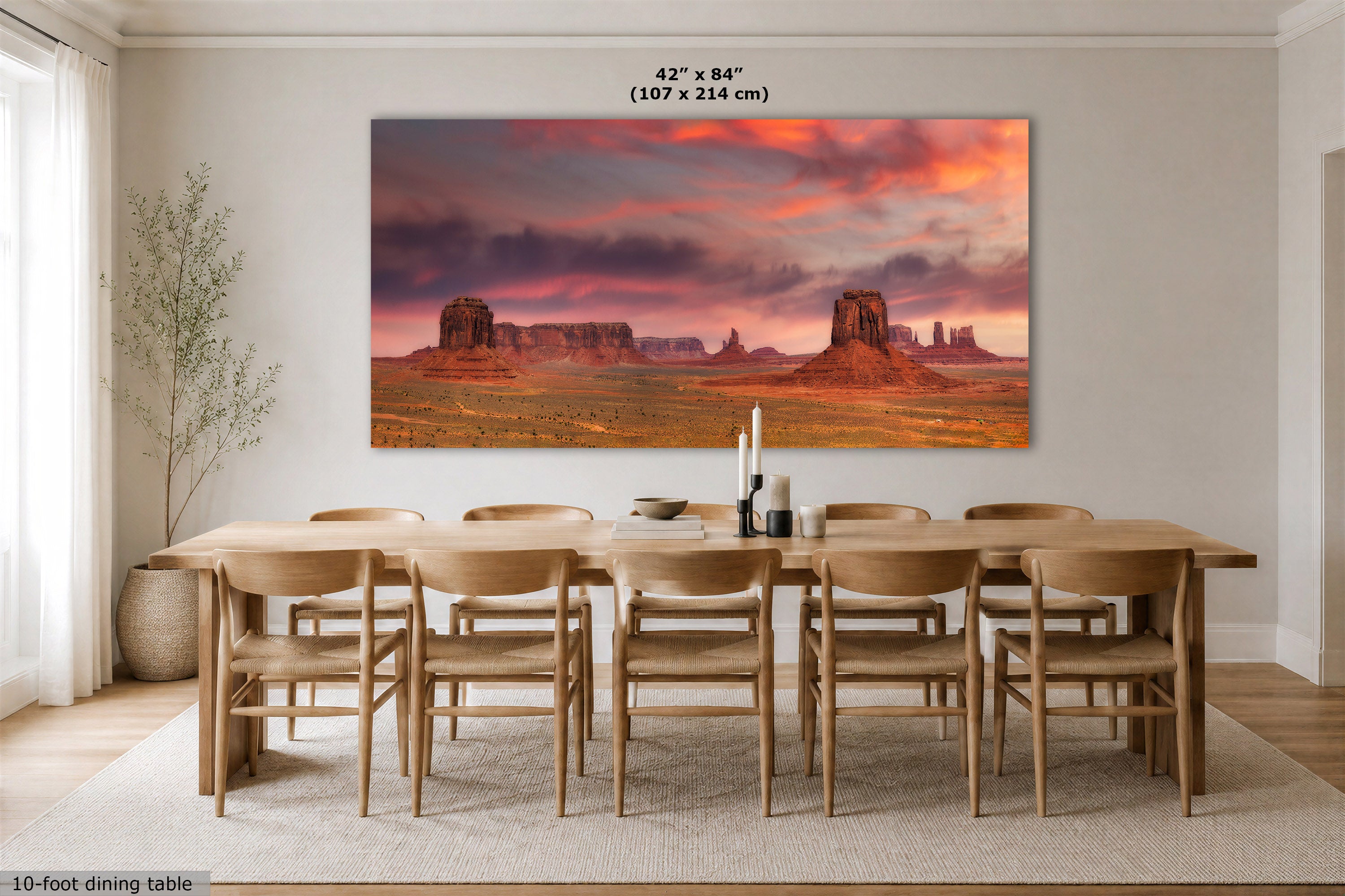 A large rectangular wall art print showing a desert landscape with rock formations under a dramatic pink and orange sky, displayed above a wooden dining table in a bright dining room. Text at the top reads 42" x 84" (107 x 214 cm), and text at the bottom left says 10-foot dining table.