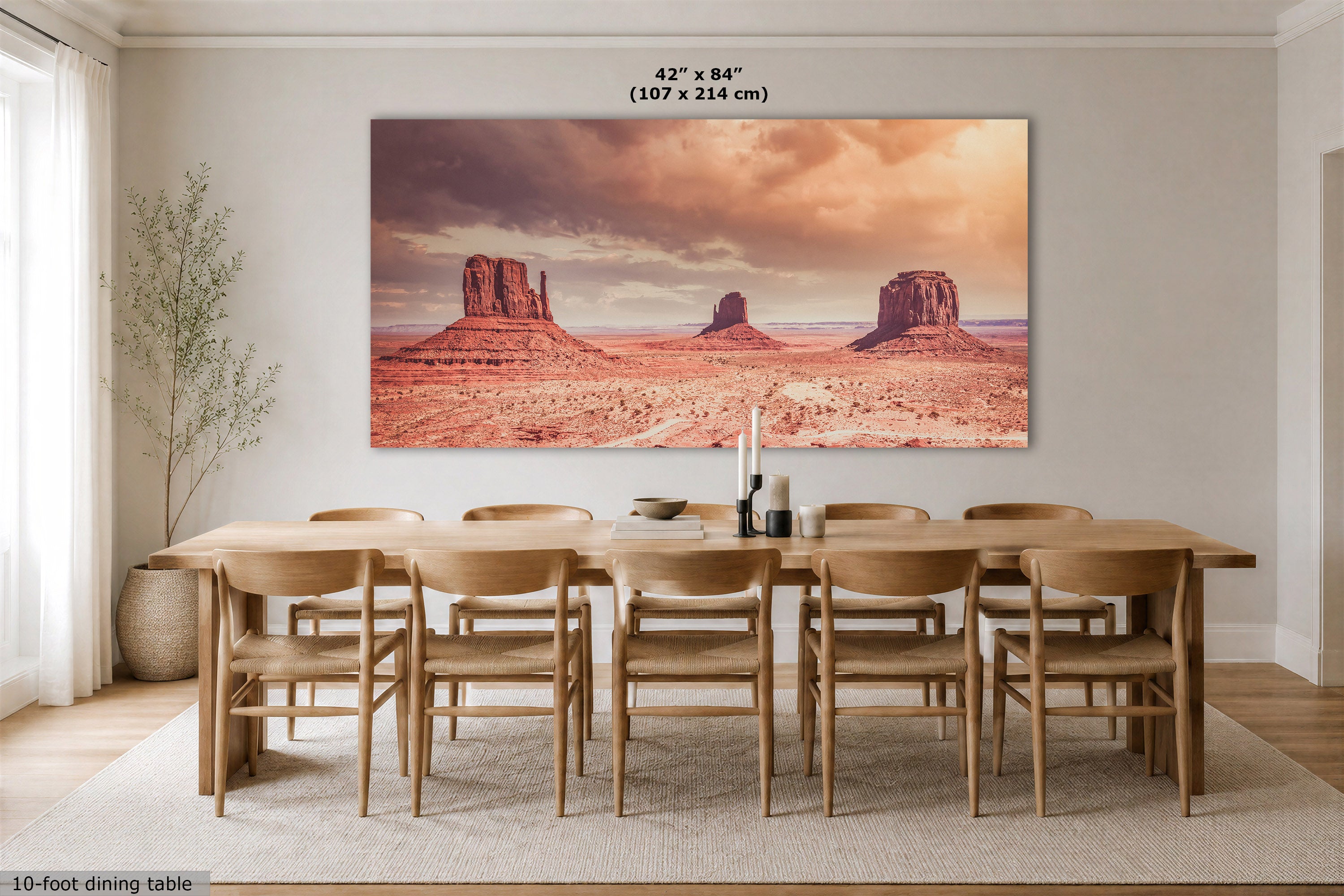 A large rectangular wall art print showing a desert landscape with rock buttes under a dramatic orange sky, displayed above a wooden dining table and chairs in a bright dining room. Text at the top reads 42" x 84" (107 x 214 cm), and text at the bottom left says 10-foot dining table.