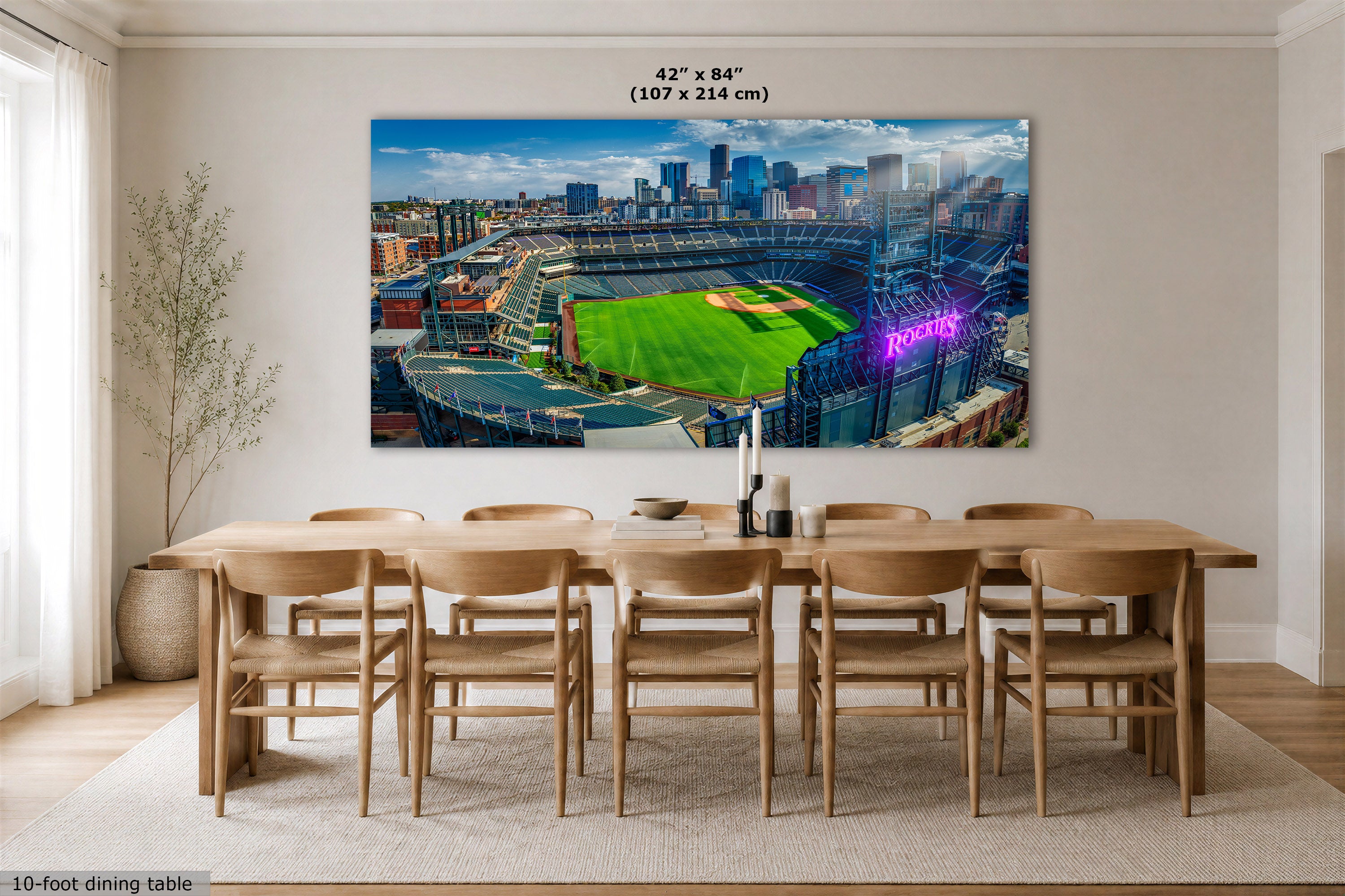A large rectangular wall art print showing the Colorado Rockies baseball stadium in Denver with a green field, blue sky, and city skyline, displayed above a wooden dining table in a bright dining room. Text at the top reads 42 x 84" (107 x 214 cm), and text at the bottom left says 10-foot dining table.